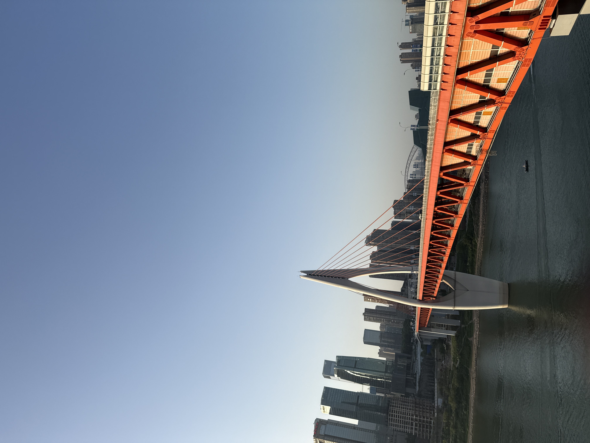 The image shows the Hongyadong Bridge, an iconic landmark in the Yuzhong District of Chongqing, China. It is a red steel arch bridge spanning a river. On either side of the river, there are multiple high-rise buildings forming a cityscape. The sky is clear and blue. The bridge's structure is prominent, with its arch design and steel support beams. Water reflects the bridge.