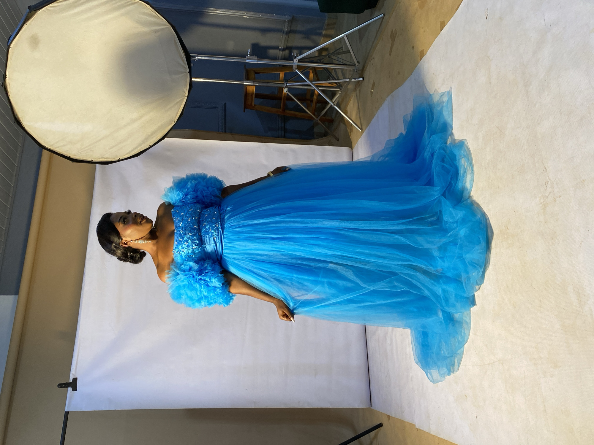 A young Black woman stands in a studio setting, her body angled towards the right of the frame. She is wearing a voluminous, bright blue tulle gown with a fitted, embellished bodice. The dress features fluffy, ruffled off-the-shoulder sleeves and a flowing skirt. Her hair is styled up, and she is wearing earrings and a necklace. The background is a seamless white backdrop, with a soft light source, likely a studio umbrella or softbox, visible in the upper left corner of the image. The lighting creates a dramatic effect, casting shadows and highlighting the texture of the dress. A metal stand with some wooden elements is visible in the background behind the woman. The scene appears to be a professional photoshoot. The overall mood is elegant and poised. There is no visible text in the image.