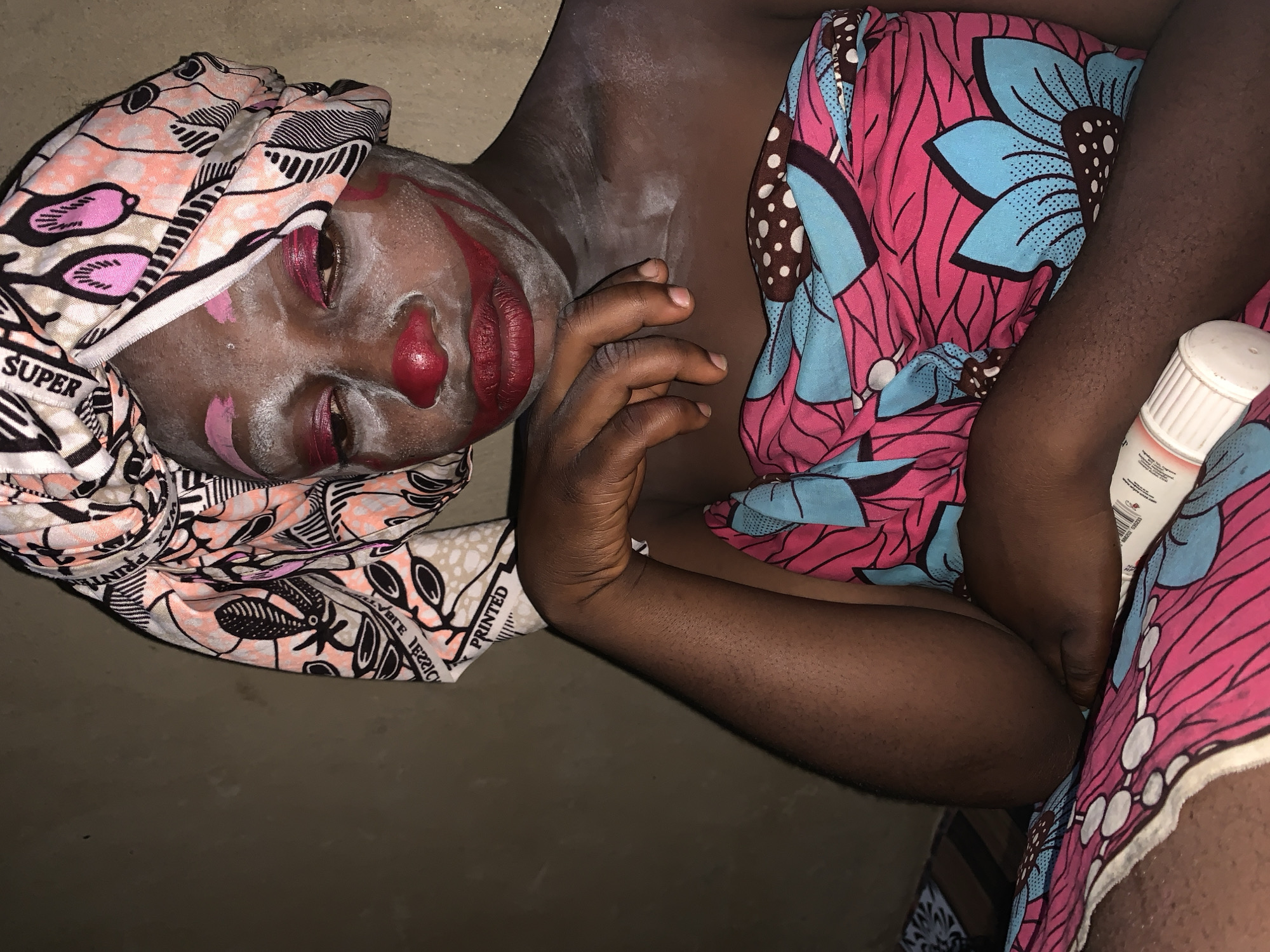 The image shows a person with clown makeup in Agban, Nigeria. The person is wearing a colorful headwrap and a patterned cloth around their body. The makeup consists of white face paint, a red nose, red lipstick, and red eyeshadow. There are also red lines painted on the face, possibly to resemble a smile. The person's left arm is resting on their leg, and their head is tilted down. A tube of what appears to be makeup or lotion is visible on the patterned cloth in the lower right corner. The background is a plain, dark wall.