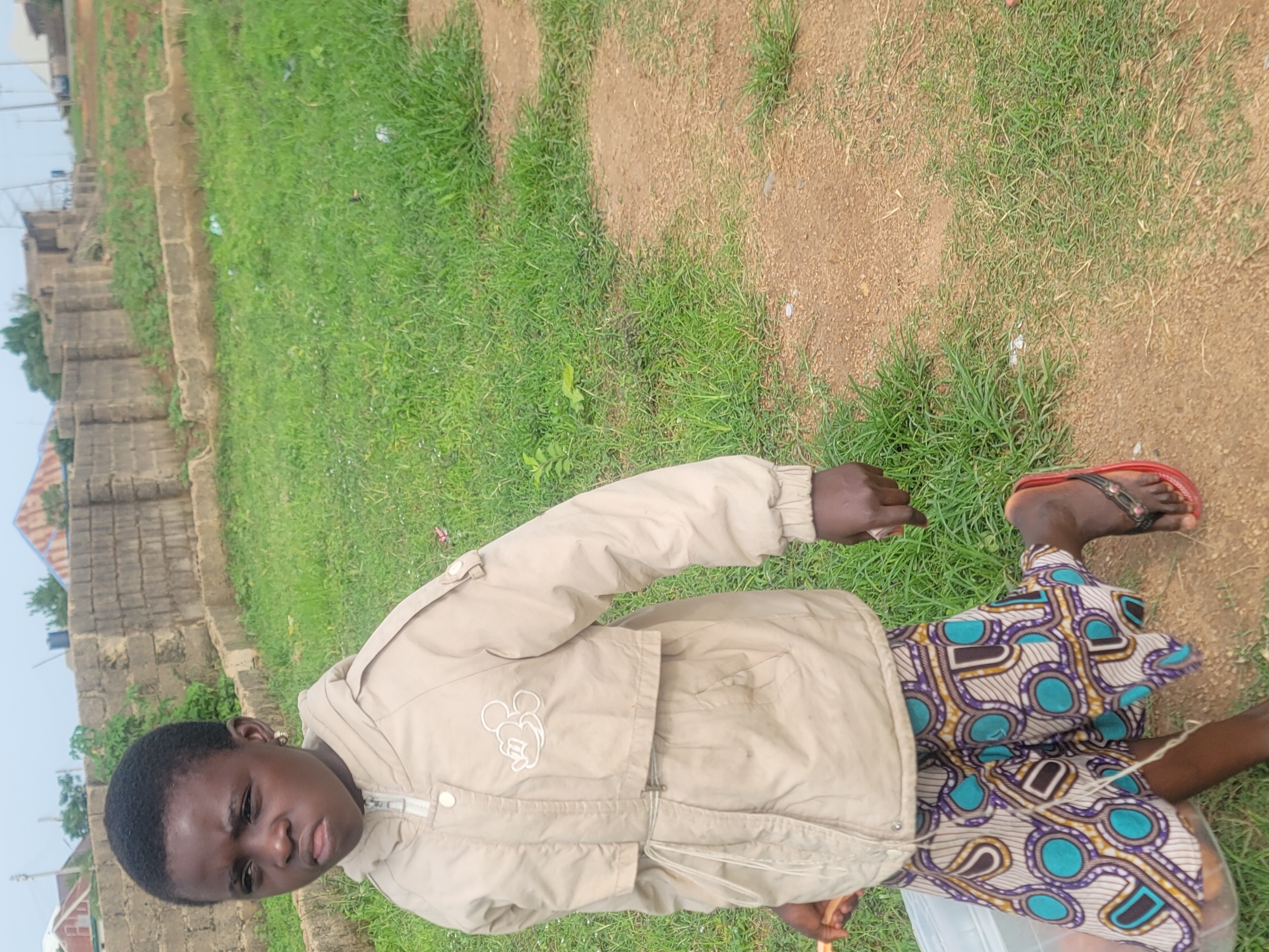 In the city of Jos, Nigeria, a young girl is captured in a medium shot, standing on a dirt path surrounded by dry grass. She is wearing a light brown hooded jacket with a Mickey Mouse emblem on the chest and a vibrantly patterned skirt. In her right hand, she holds a light brown object, possibly a stick, and a clear plastic container is visible in her left hand. The background consists of a low, rough-hewn stone wall and more dry grass, with a glimpse of buildings and structures further in the distance under a hazy sky. The scene appears to be an outdoor, semi-urban environment.
