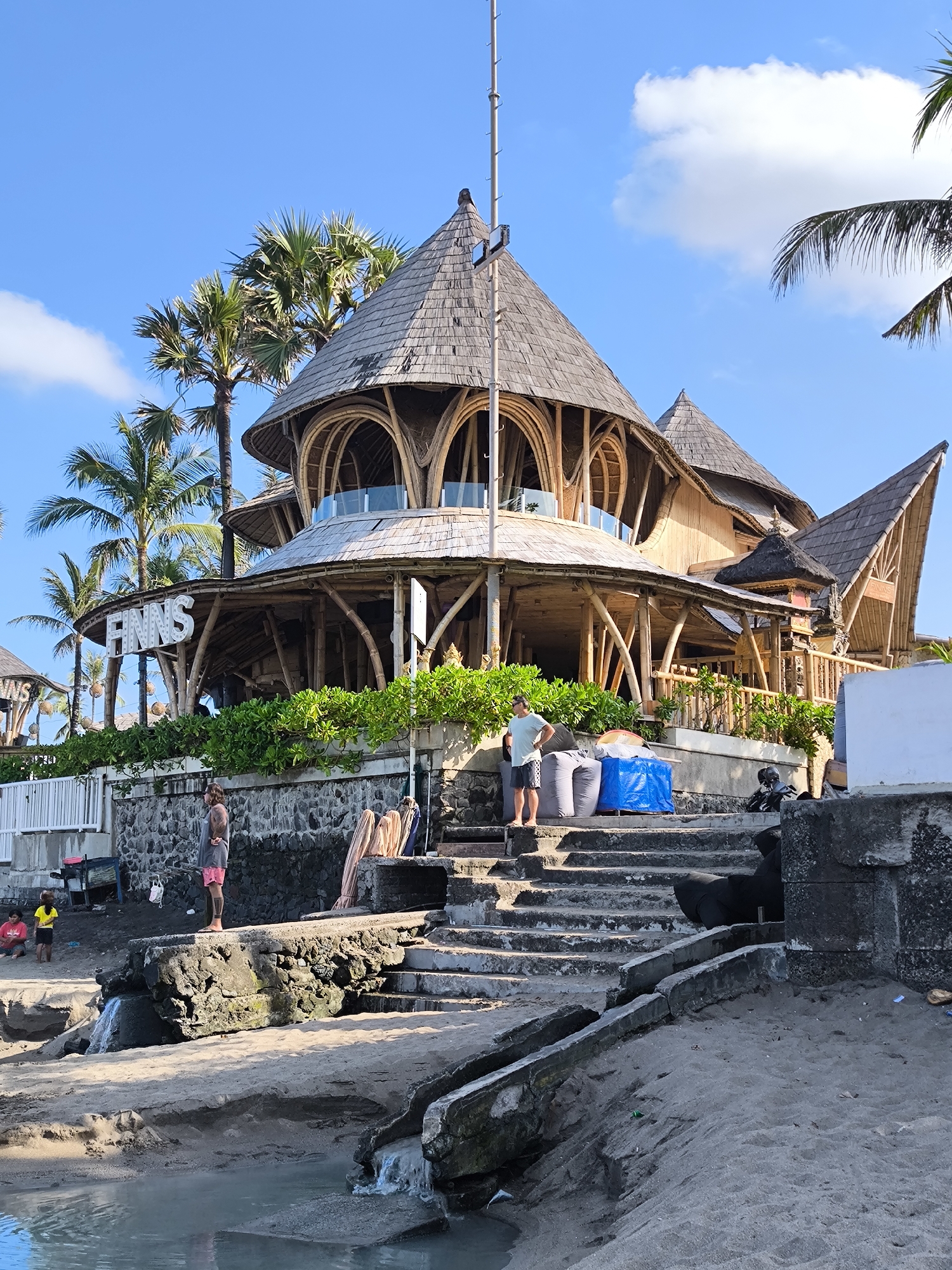 The image shows a scene in Tibubeneng, Indonesia, featuring a multi-story building made of bamboo and wood, identified by a sign reading "FINNS." The building has a distinct layered, conical roof and an open-air design. Lush greenery lines the base of the structure.
In the foreground, a set of concrete steps lead down to a beach area with dark sand and standing water. Two people are visible on a raised concrete platform adjacent to the steps. One person is standing on the platform, another person wearing shorts and a sleeveless shirt with visible tattoos standing on the other side of the stairway, looking out towards the water. Further back, a man wearing a t-shirt and shorts stands on the stairway near the base of the building. A few other people and what may be children are further to the left, near the shore.
Large, dark bags are visible near the man standing on the stairs, covered in a blue tarp. Debris and trash are scattered along the beach and steps, indicating possible pollution. Palm trees and a clear blue sky with scattered clouds complete the background.