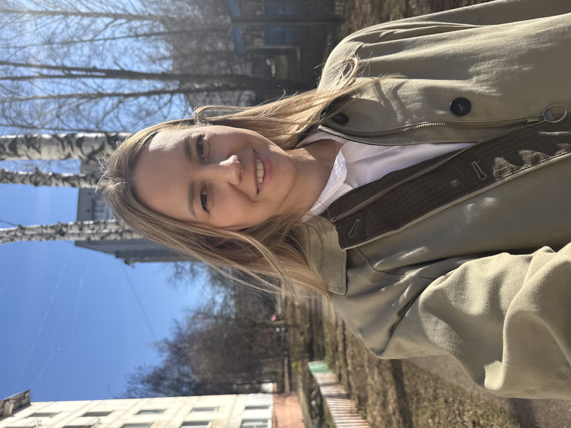A young woman is depicted in an outdoor setting under bright daylight, likely in Chaikovsky, Russia, during late winter or early spring. The image is oriented sideways. She has light blonde hair and is smiling broadly at the camera, conveying a cheerful and relaxed expression. She wears a light-colored jacket, possibly olive green or beige, which features a zipper closure and a button detail on the lapel, over a white collared shirt.
The background features a clear, vivid blue sky. Bare deciduous trees, including several distinctive birch trees with white bark, are prominent, indicating the season. Multiple buildings, one a light-colored, multi-story residential structure with visible windows, are also part of the background. The ground beneath the trees shows dry grass and patches of earth. Strong, directional sunlight illuminates the scene, casting clear shadows. No visible text is present in the image.