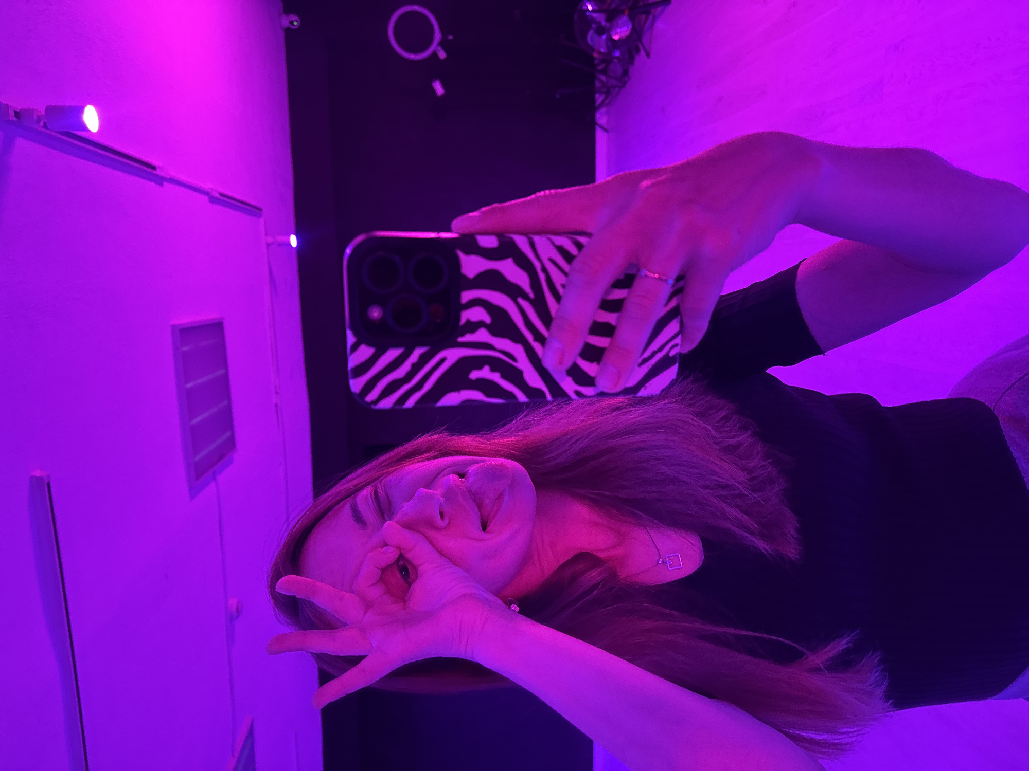 A woman is taking a selfie in what appears to be an interior space, judging by the presence of walls and lighting fixtures. The walls are illuminated with purple light. She is holding a smartphone with a zebra-striped case, which is obscuring part of her face. Her hair is brown, and she is wearing a black top. She is making an "OK" gesture with one hand. The location is specified as Chaikovsky, Russia.