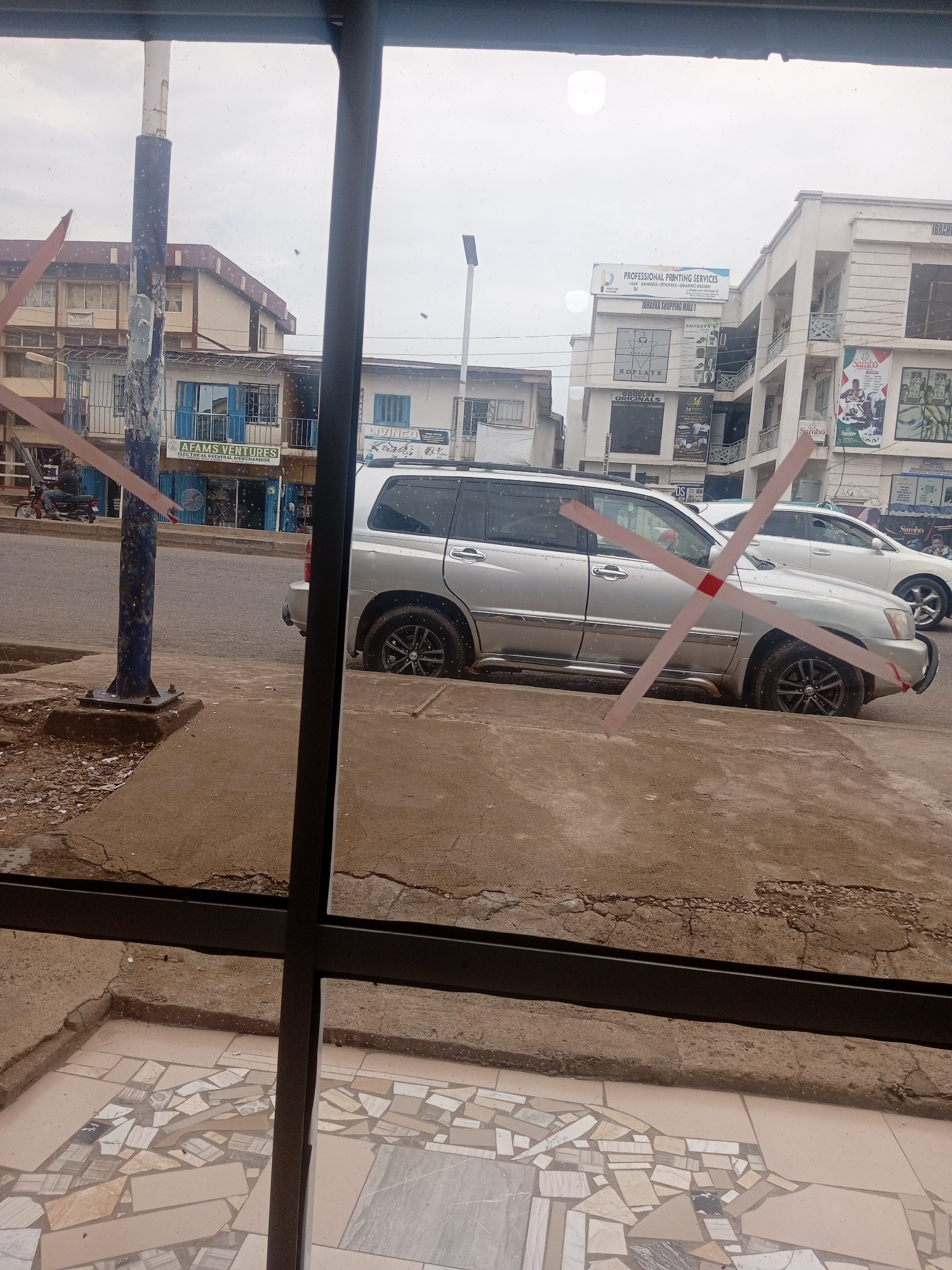 The image captures a street scene in Sardauna, Nigeria, viewed through a window. In the foreground, a portion of a tiled floor and a concrete walkway are visible, partially obscured by a dark vertical frame. A silver SUV is parked on the side of a paved road. Behind the SUV, a white car is also visible. Several commercial buildings line the street, with signs indicating businesses such as "AFAMS VENTURES ELECTRICAL/GENERAL MERCHANDISE" and "PROFESSIONAL PRINTING SERVICES IBARRKA SHOPPING MALL". The sky appears overcast. Red and white tape is placed in an 'X' formation across the foreground of the image, obscuring parts of the scene.