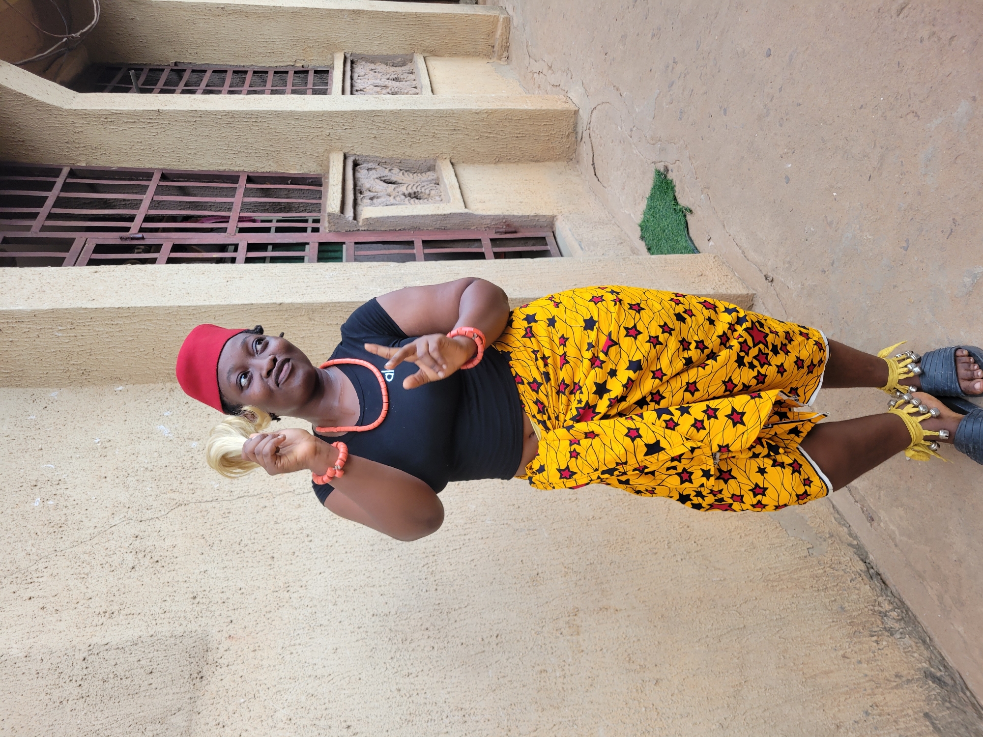 A woman stands on a sandy, textured surface in front of a multi-story building in Onitsha, Nigeria. The building has a weathered, tan-colored facade with rectangular window openings protected by dark horizontal bars. Artificial green turf is visible in a recessed area near the building's base. The woman wears a red cap, a black top, a coral beaded necklace and bracelet, and yellow shorts with a red star pattern. She holds a bundle of light blonde hair in her left hand and gestures with her right hand. Her feet are adorned with silver bells tied to yellow fabric and she wears blue sandals.