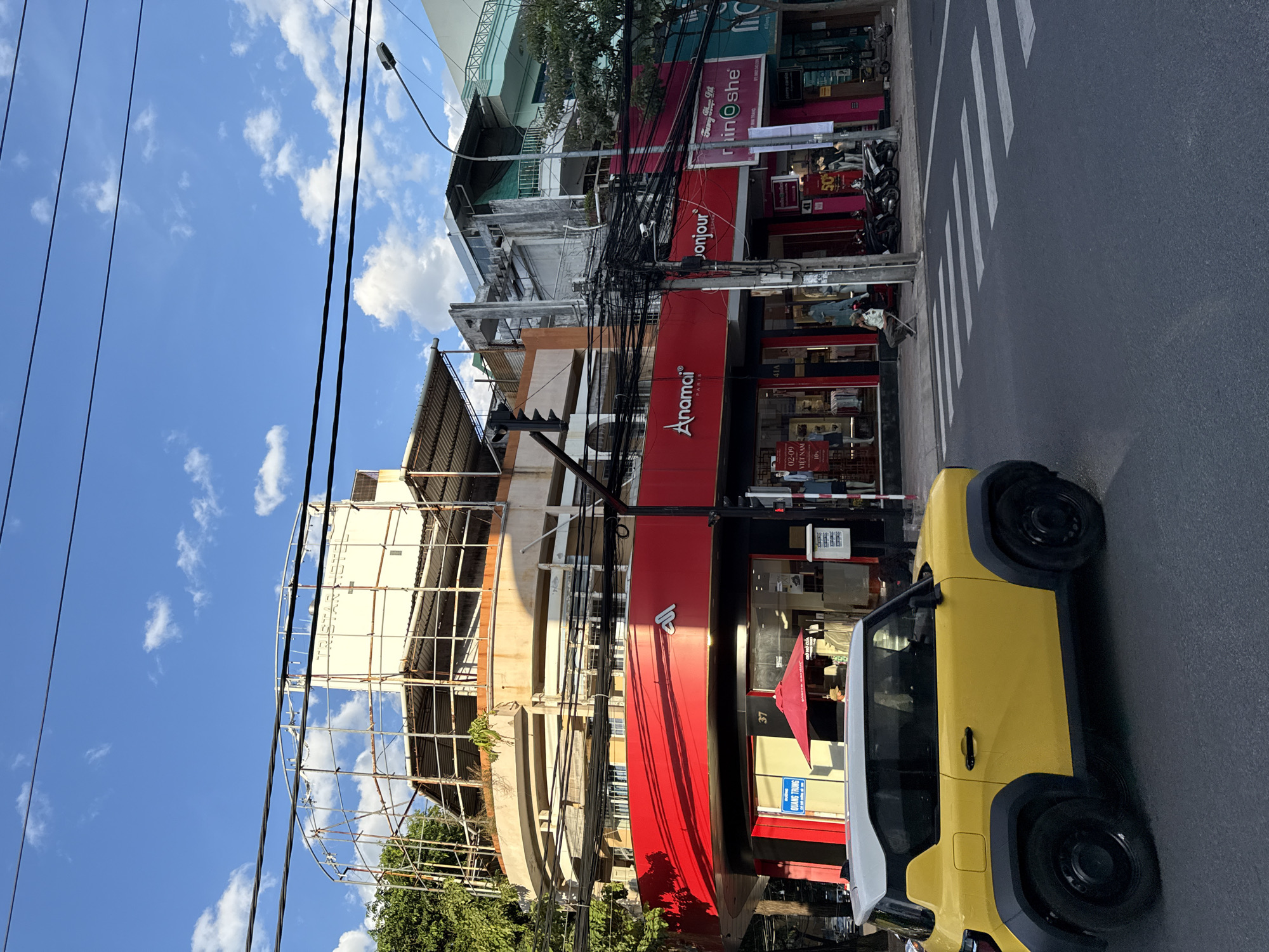 The image depicts a street scene in Nha Trang, Vietnam, on a sunny day with a blue sky and scattered clouds. In the foreground, a bright yellow and white compact car is parked on the left side of the frame, partially obscuring a pedestrian crossing. Across the street, a row of buildings lines the sidewalk. The buildings feature storefronts with prominent signage, including "Anamci PARIS", "Bonjour", and "riin oshe". Some of the shops display clothing items. Above the storefronts, the buildings are older, with some appearing to be under renovation or in a state of disrepair, as indicated by scaffolding on the top levels. Numerous electrical wires are strung across the street, creating a dense network overhead. A few motorcycles are parked near the sidewalk. A person is seated on a chair outside one of the shops. A red umbrella is visible above the entrance of a store with the number "37" displayed. The overall atmosphere is that of a bustling urban environment.