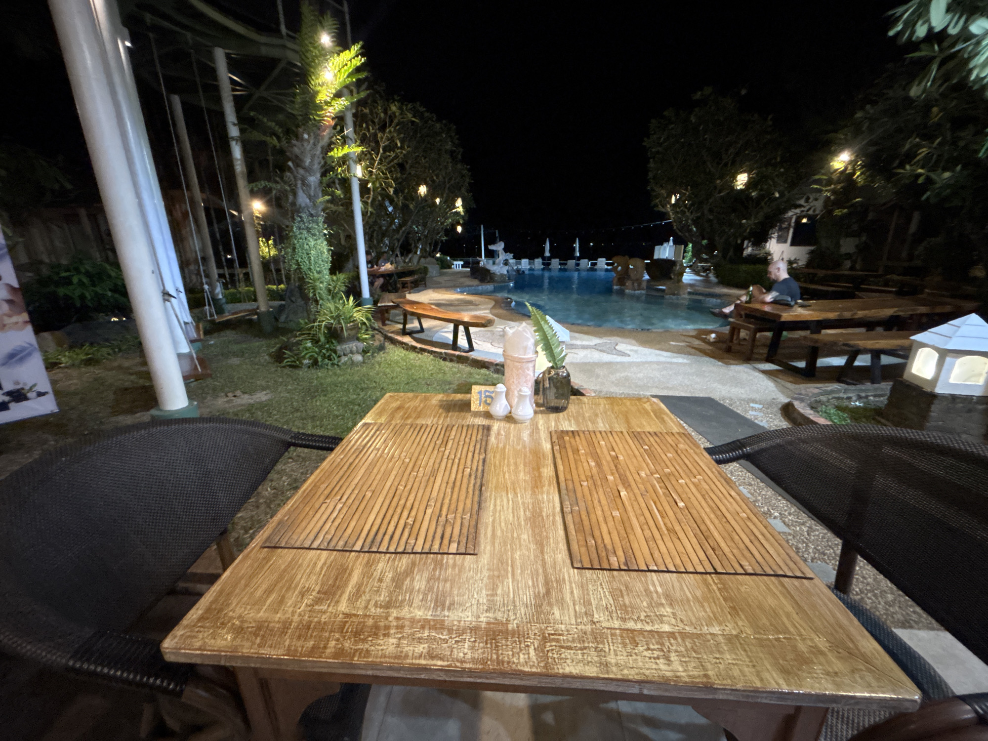 In the city of Chalong, Thailand, at night, a wooden table is set for dining outdoors. The table is situated on a paved area, with two black woven chairs on either side. Two bamboo placemats are placed on the table, and on one of them, a table number "15" is visible, along with two small white salt and pepper shakers and a small vase with a fern. In the background, a swimming pool glows under the night sky, surrounded by trees and lounge chairs. A person is seated at a picnic table by the pool, holding a bottle. Swings are visible to the left of the frame, and decorative lights are strung throughout the area, illuminating the scene.