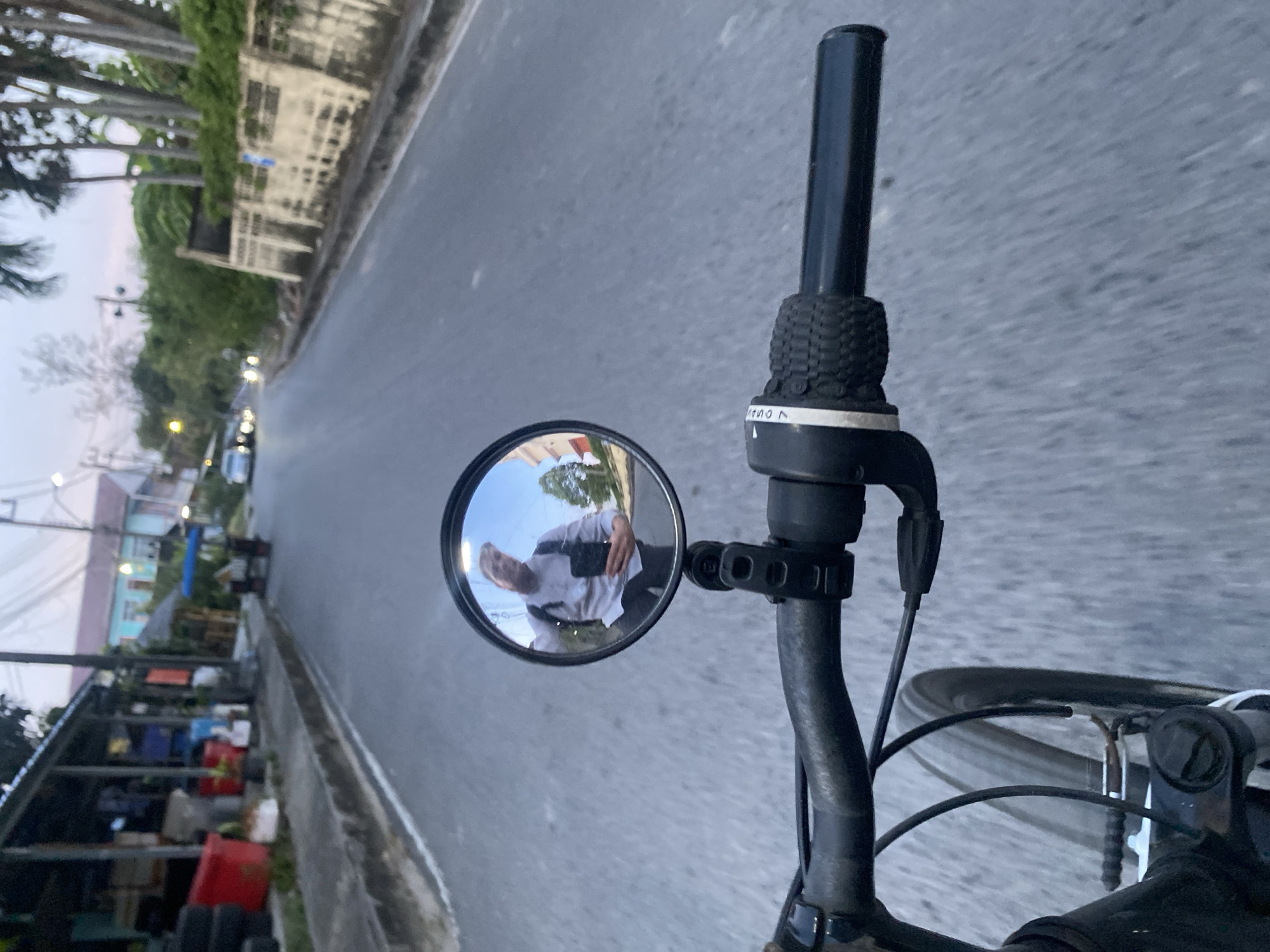 The image shows a first-person view from a bicycle on a road in Choeng Thale, Thailand. A circular mirror attached to the handlebar reflects the rider, who is wearing a white shirt and appears to be taking a photo with their phone. The road is asphalt and stretches ahead, flanked by buildings and trees on either side. One building is partially visible behind an adjacent covered structure; it's blue and red with some scaffolding. Light poles run alongside the road. The bicycle handlebars, grips, and front wheel are visible in the foreground. A 7-speed indicator is on the handlebar.