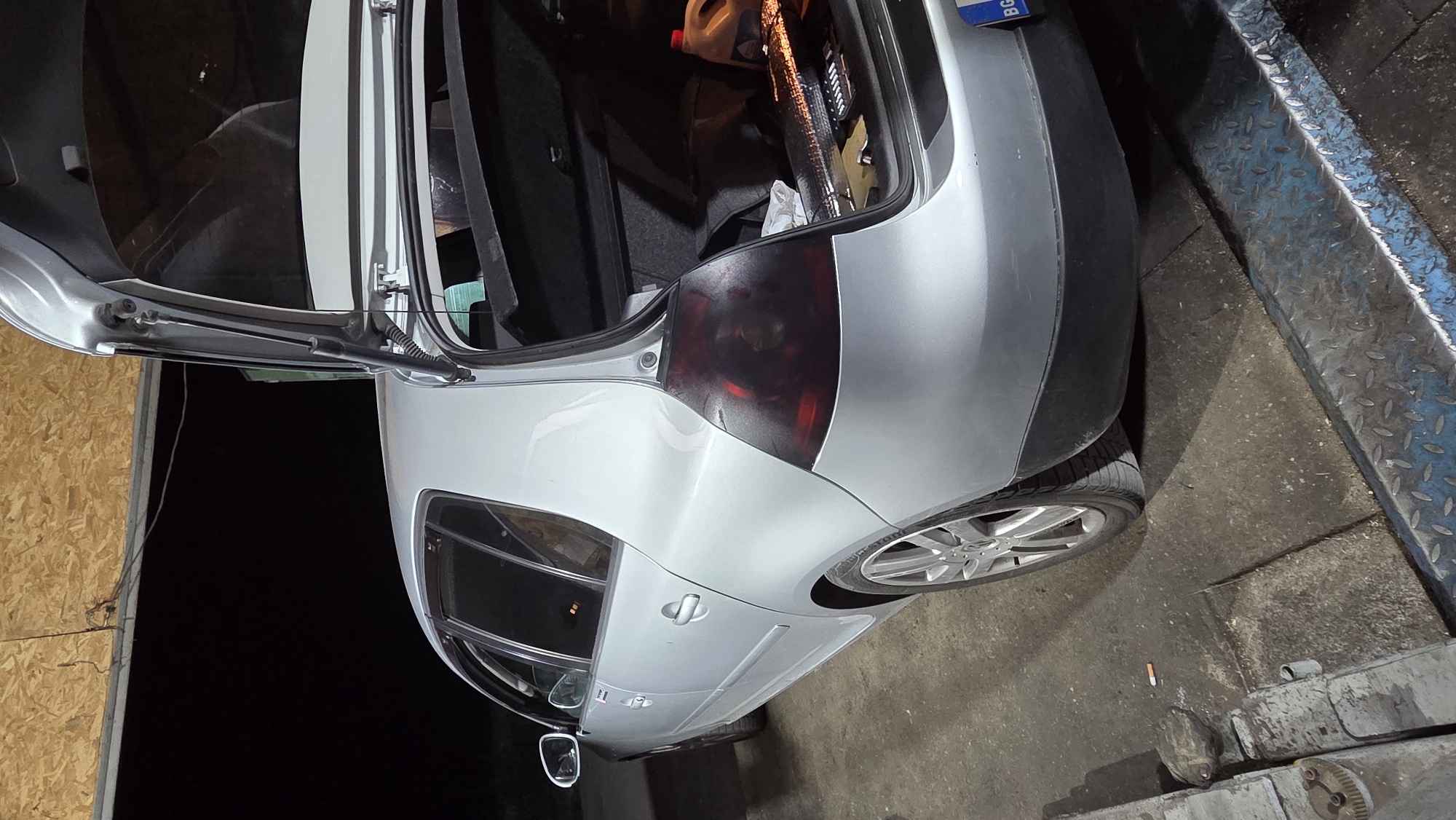 In an indoor garage, a silver hatchback car is parked. The rear hatch is open, revealing the trunk filled with various items including a red plastic jug, a reflective material, and other unidentifiable objects. The car is positioned on a platform with a metal diamond plate surface. The floor is concrete and appears worn. A discarded cigarette butt is visible on the ground near the front wheel. The walls of the garage are made of wood paneling. The lighting suggests it is nighttime. The license plate of the car is partially visible and reads "BG".