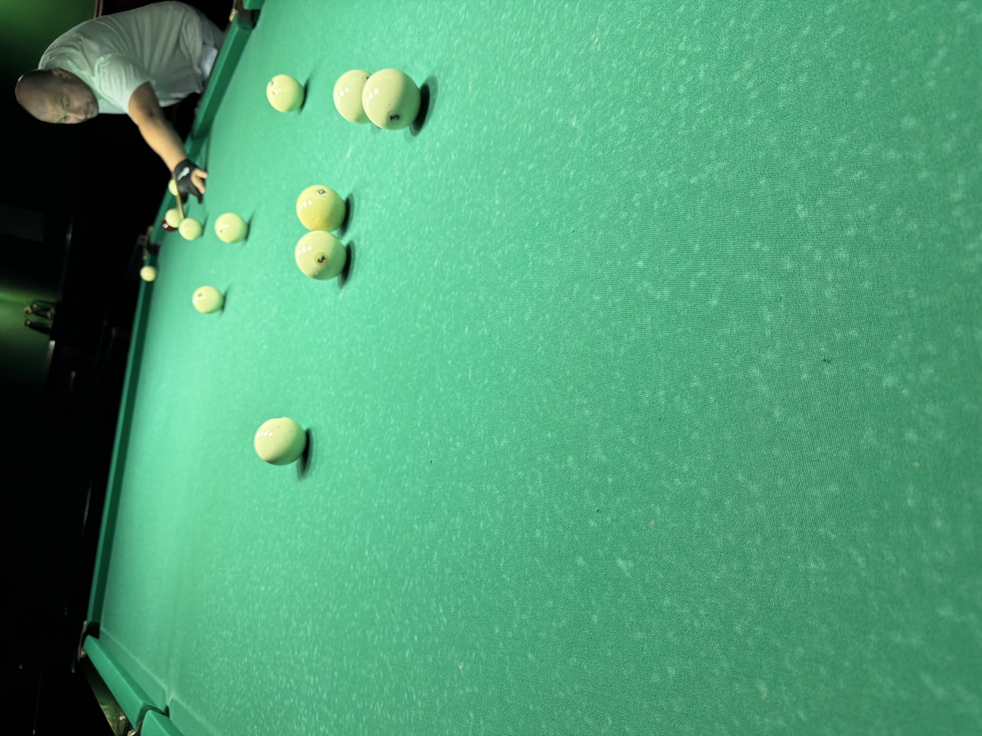 The image depicts a billiard table with several balls scattered across its green felt surface. A man, appearing to be of bald head, is visible in the background, positioned behind the table and in the process of playing. He is wearing a light-colored t-shirt and is in the act of striking a billiard ball with a cue stick. The environment appears to be an indoor setting, possibly a billiard hall, with a dark background and a green illuminated ceiling. The perspective of the shot is from a low angle, looking up at the billiard table and the player. There is no indication of the location being in Strășeni, Moldova, as this information is not visually present in the image.