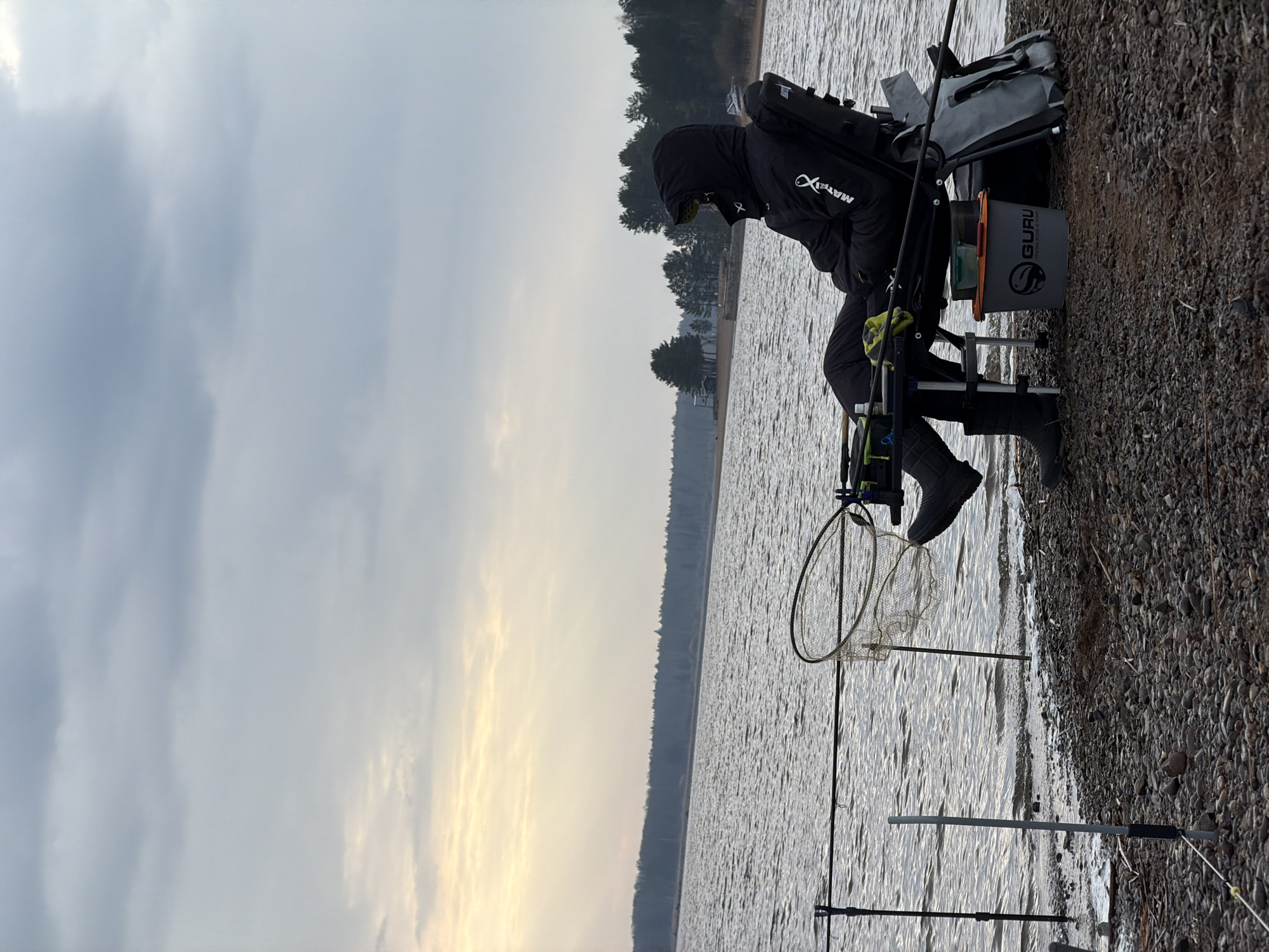 An individual is captured in an outdoor setting, engaged in fishing by a large body of water. The person, viewed from the side, is seated in a specialized fishing chair on a pebbly bank. They are dressed in warm clothing, including a black hooded jacket with "MATRIX" and a fish logo visible on the sleeve and back. Black boots are also visible on their feet.
In front of the person, fishing rods are set up, extending into the water. A fishing landing net stands upright near the water's edge. Beside the person's chair, there's a grey fishing bucket or tackle container with the text "GURU KNOWLEDGE IS POWER" printed on it, along with a fish motif. A grey bag is also strapped to the back of the fishing chair.
The scene depicts a calm, overcast day, possibly late afternoon or early morning, as indicated by a slightly brighter, hazy band on the distant horizon. The water appears slightly choppy with ripples. The background features a forested shoreline with numerous trees, suggesting a lake or wide river, consistent with the location context of Chaikovsky, Russia. The overall atmosphere is quiet and focused on the fishing activity.