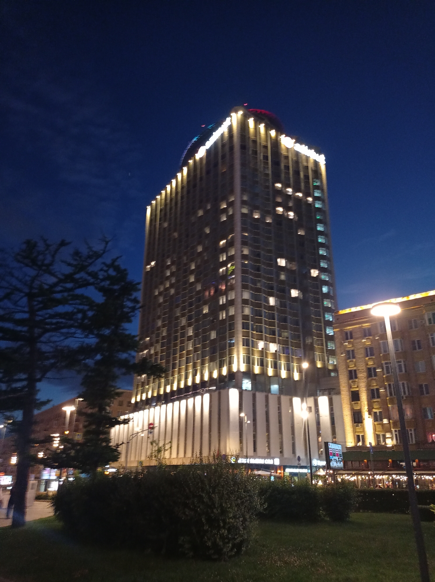 The image depicts a nighttime scene in Moscow, Russia, focusing on the Arbat High-Rise Building, also known as the Ministry of Foreign Affairs Building. The imposing skyscraper, illuminated by warm lights along its edges and numerous interior lights glowing from its windows, dominates the composition. The Arbat High-Rise is characterized by its distinct Stalinist Empire style architecture. To the right of the Arbat High-Rise, a shorter, older brick building with arched windows is visible, also lit from within and along its upper facade. In the foreground, a dark, manicured lawn is interspersed with dense green bushes and a solitary, silhouetted tree. Streetlights, one a tall modern lamp post, cast a soft glow on the scene, enhancing the nocturnal ambiance. The sky above is a deep twilight blue, with faint wisps of clouds. The overall impression is one of urban nighttime grandeur and historical architectural presence in the heart of Moscow.