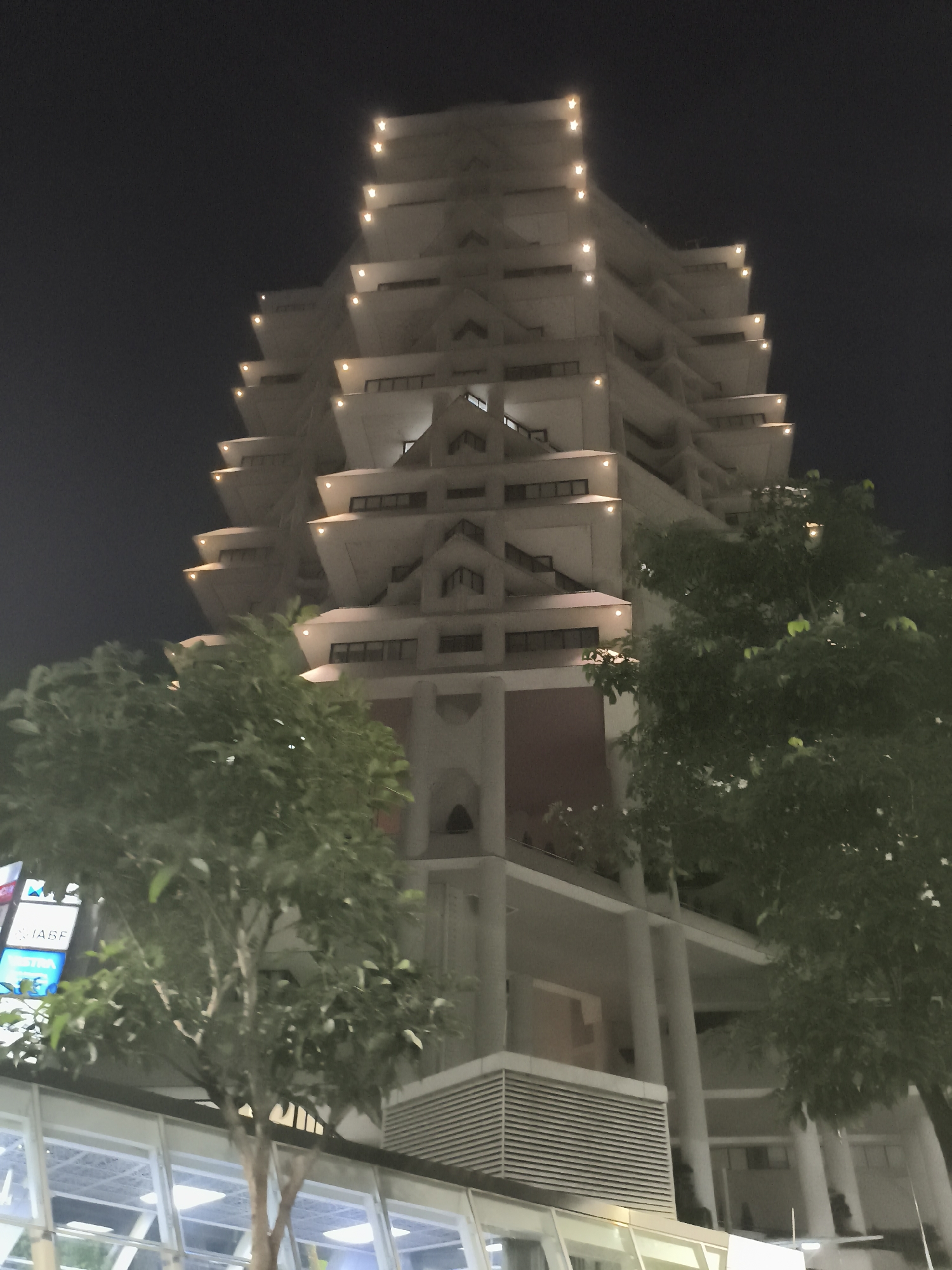 The image shows a night view of the central library of the University of Indonesia, located in South Jakarta, Indonesia. The multi-tiered building is predominantly white, with a unique stepped design reminiscent of a pagoda. Each tier is illuminated with lights along its edges, creating a distinct pattern against the dark sky. The building is partially obscured by trees in the foreground, framing the structure. In the lower-left corner, a portion of a glass-walled structure with signs is visible.