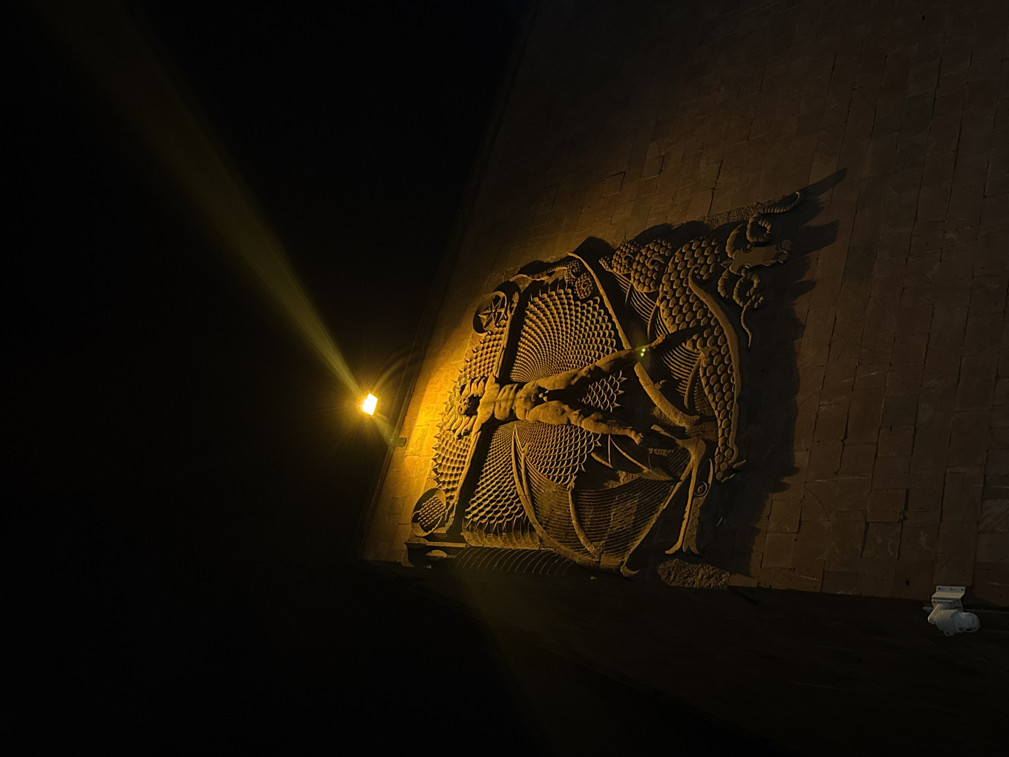 A low-angle, long shot captures a carved relief illuminated by a light at night. The relief, which appears to be made of stone, is fixed on a brick wall. The relief is an intricate design with a human figure that is encircled by what appears to be a phoenix. The entire design has an ancient and possibly Eastern Asian appearance.
The scene is darkly lit, with the wall and the relief cast in deep shadows. The source of light shines from the left onto the relief, highlighting the detail of the carving. A beam of light is visible in the upper left of the photo, adding to the contrast. A small white object sits on the bottom right corner of the brick wall, near the sculpture.