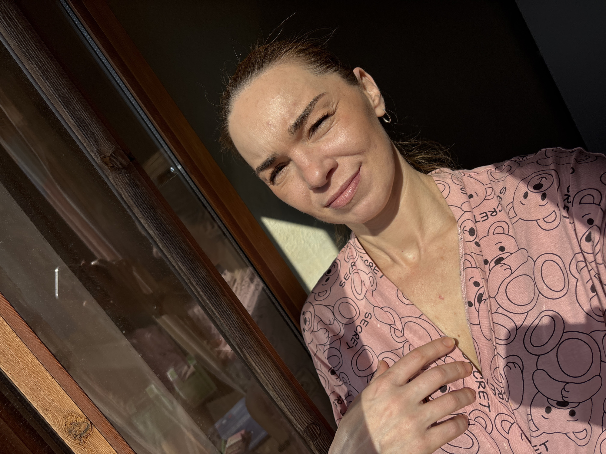 A woman is pictured from the chest up, looking to her right with her eyes squinted due to the sunlight. She is wearing a pink pajama top with a pattern of cartoon teddy bears and the word "SECRET" repeated. Her right hand is resting on her chest. The background is dominated by a slanted wooden-framed window with sunlight streaming through, casting a shadow on the right side of her face and neck. The window appears to look out onto an outdoor scene, possibly a city, though details are not clear.