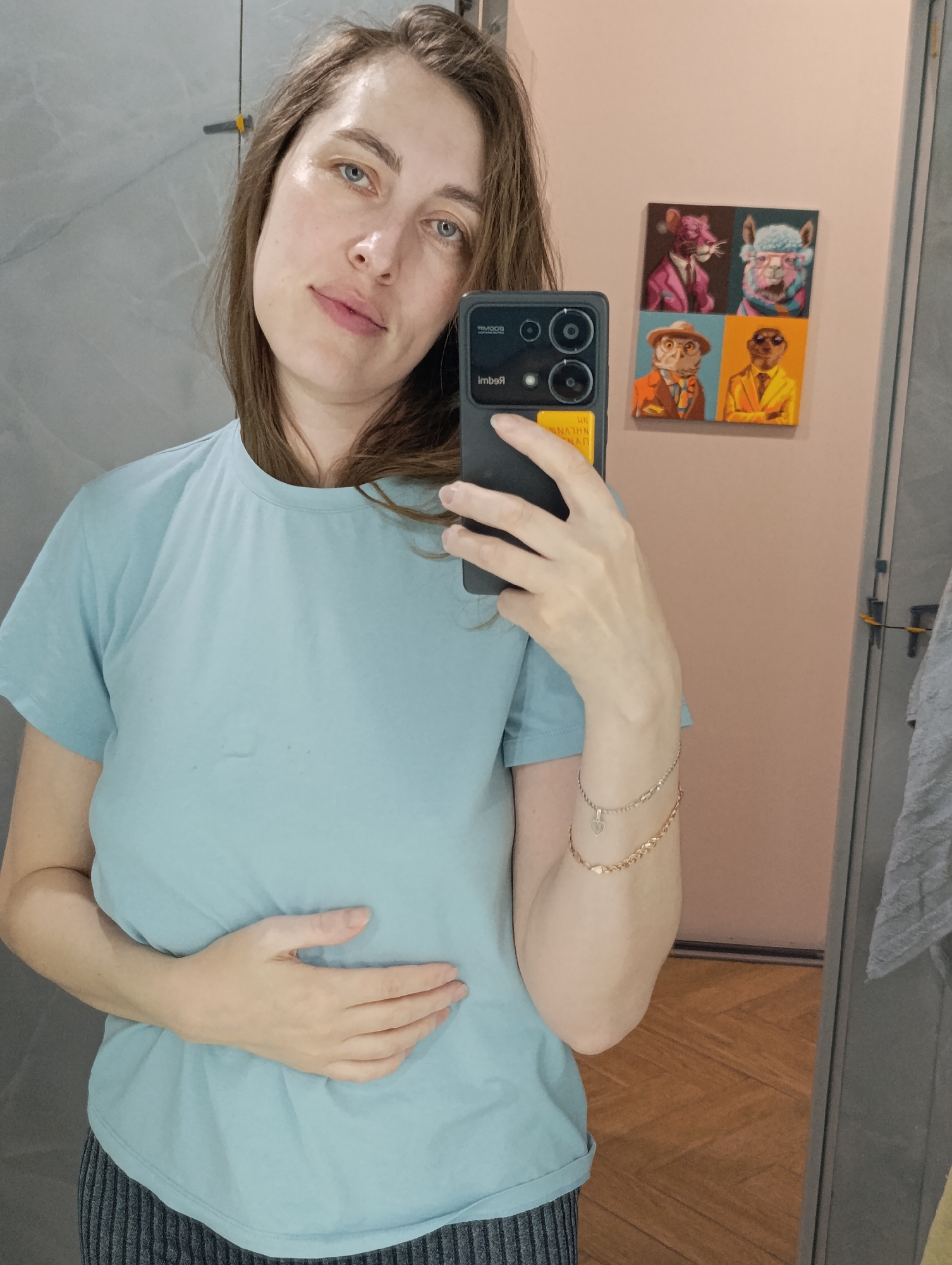 A fair-skinned woman with light brown hair and blue eyes takes a selfie in a bathroom. She's wearing a light blue T-shirt and what appears to be gray striped pajama bottoms. Her left hand is resting on her stomach. Two gold-toned bracelets adorn her right wrist. She is holding a black smartphone. Behind her, on a light pink wall, hangs a square artwork featuring four anthropomorphic animal portraits. The location could be Chaikovsky, Russia. The surrounding environment includes marble-effect wall tiles and a wooden floor. A towel hangs on the right side of the mirror.