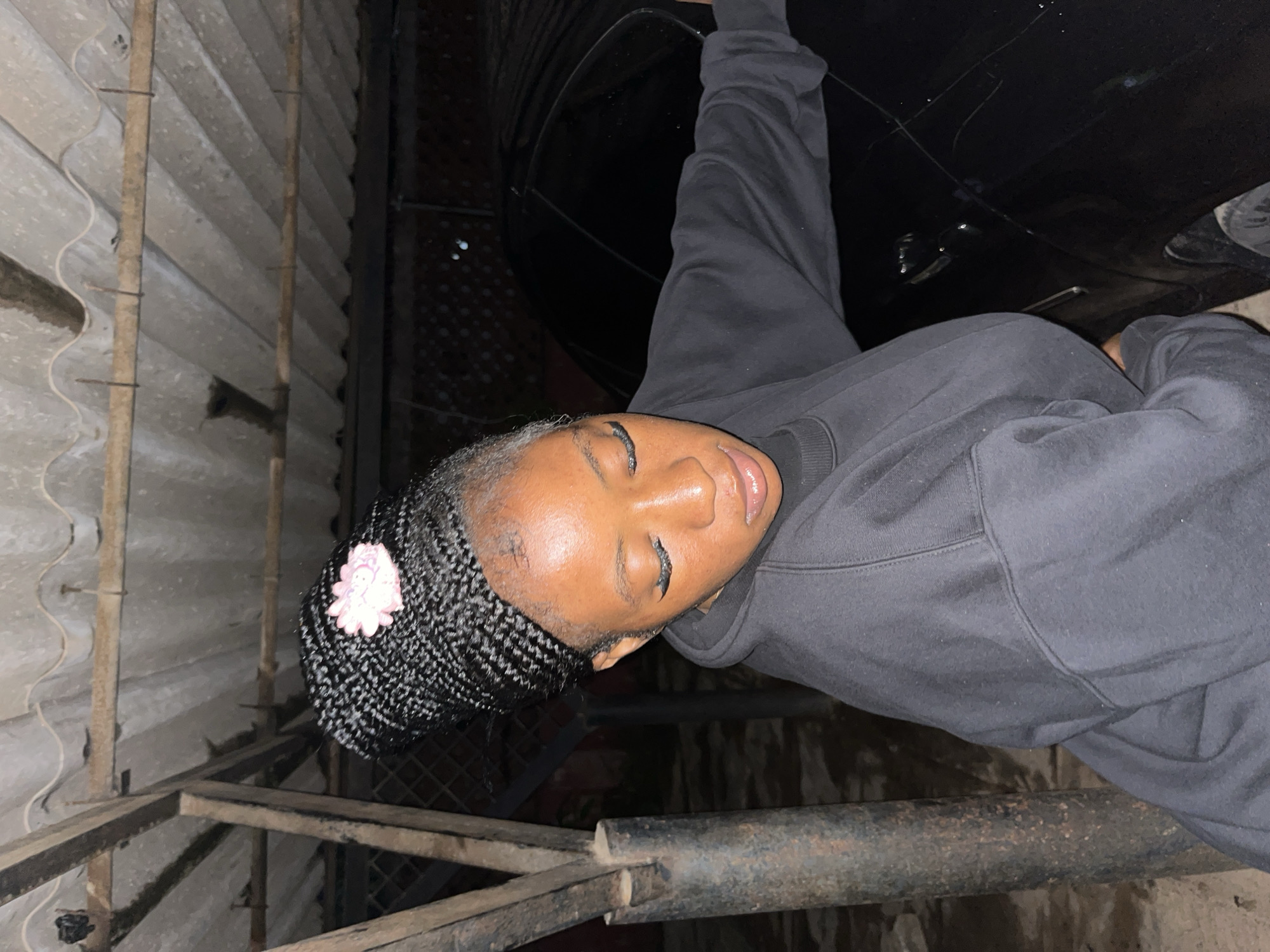 In the city of Jos, Nigeria, an individual with braided hair adorned with a pink flower is captured in a low-angle, medium shot. They are wearing a black sweatshirt and are leaning against a black car. The background features a corrugated metal roof supported by wooden beams and a thick, rusty metal pole, suggesting an outdoor, possibly informal, setting like a carport or workshop. The lighting is dim, indicative of nighttime or low-light conditions.