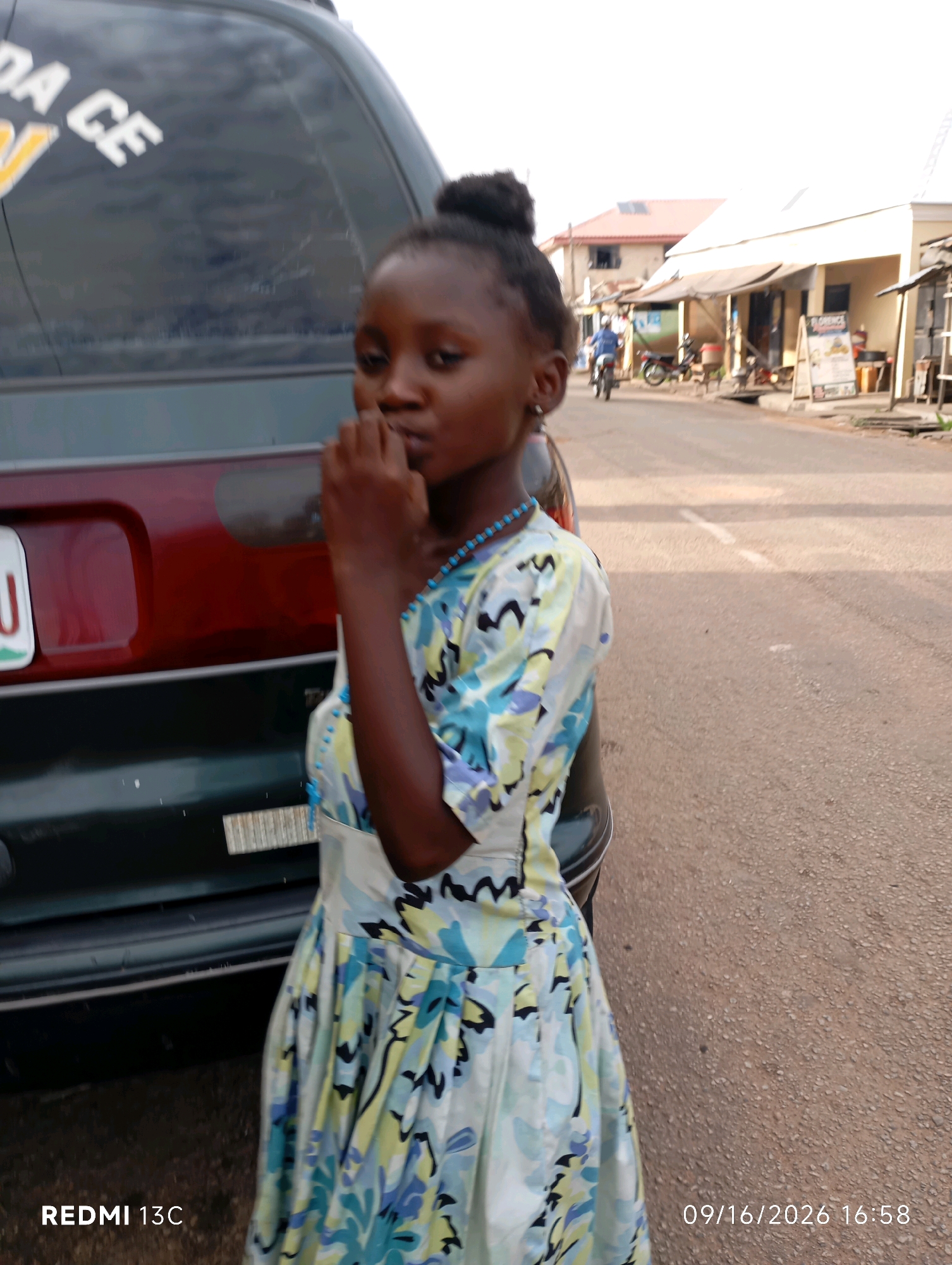 A young Black female stands next to a dark colored vehicle. The vehicle has a bumper sticker with white lettering that reads "DA CE". The girl is wearing a blue, yellow, and white patterned dress and a blue beaded necklace. Her hair is pulled up into a bun on top of her head. She is holding her hand to her mouth. The background shows a street with buildings and a few motorcycles. The time stamp at the bottom right of the image reads "09/16/2026 16:58". The location identified is Shendam, Nigeria.