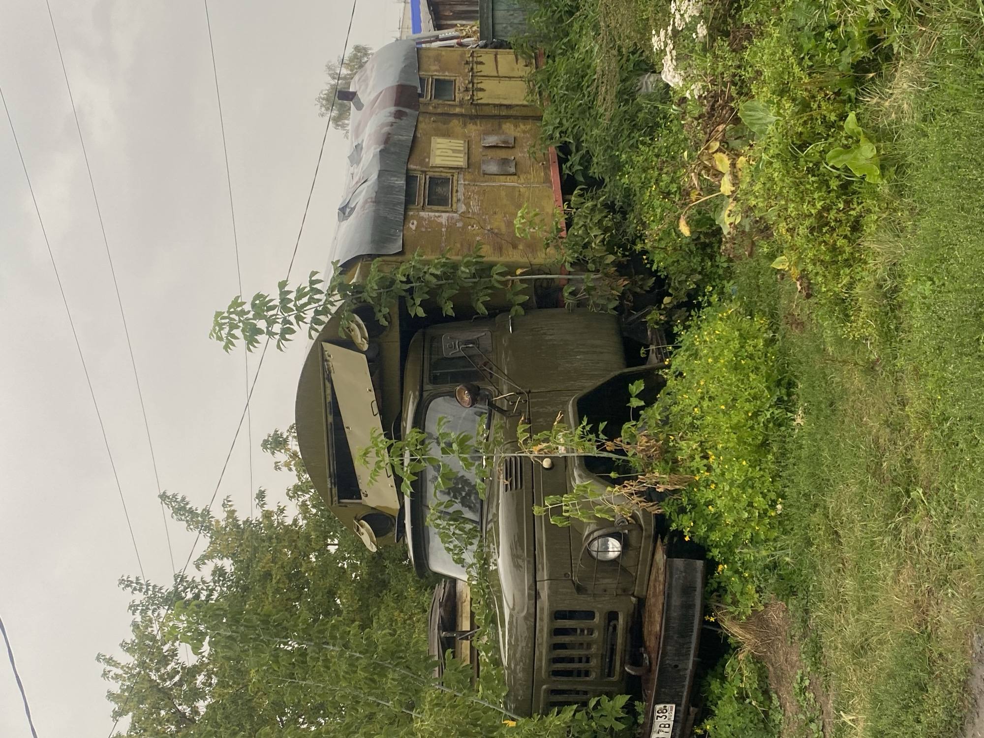 An overgrown, abandoned military truck, painted olive drab, is parked amongst dense green foliage under a cloudy sky. The truck has a modified living quarter attached to its rear, painted a faded yellow with red trim. This structure has several small windows and a corrugated metal roof. Vines and weeds have crept up the side of the truck and are beginning to cover the living quarters. The license plate on the front of the truck reads "7B 38 RUS". The scene appears to be in a rural or industrial area on the outskirts of Novosibirsk, Russia. There are no people visible in the image, and no actions are taking place.