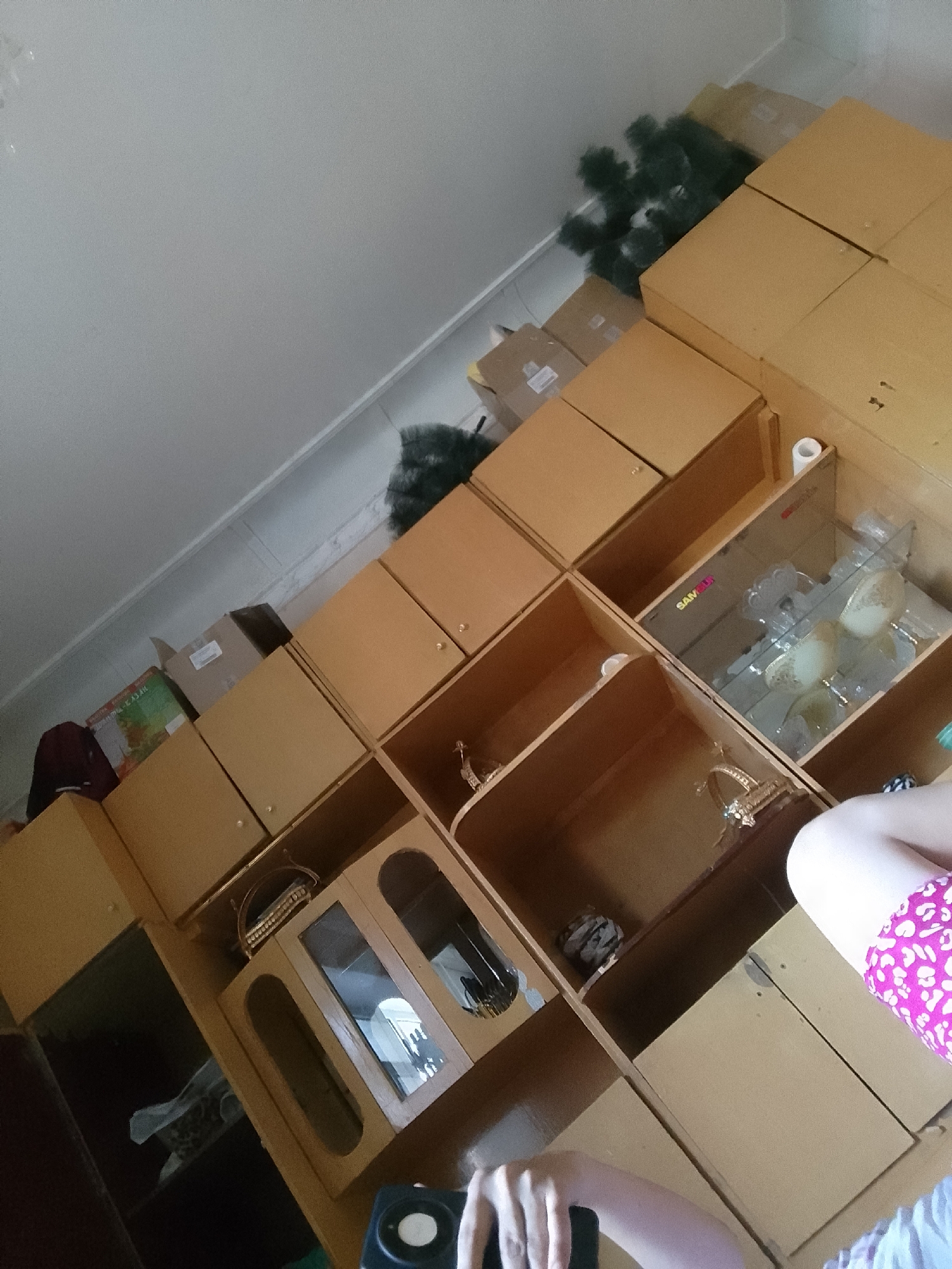 The image depicts an indoor scene with a large amount of cardboard boxes stacked and arranged in what appears to be a storage or organization area. Some of the boxes are open, revealing their contents or the inside of the storage units. A disassembled artificial Christmas tree is visible among the boxes, suggesting preparation for or cleanup after a holiday. The shelving units are made of a light-colored wood, and some compartments contain decorative items, including a gold-colored ornate stand and glass or ceramic decorative pieces. A person's leg is visible on the right side of the image, wearing pink and white patterned shorts. Another person's hand is partially visible in the foreground, holding what appears to be a black electronic device with a white button. The overall environment seems to be a room in a residence or a storage facility. The location is identified as Taxtakópir, Uzbekistan, though no specific city landmarks or contextual details are present within the image itself to confirm this.