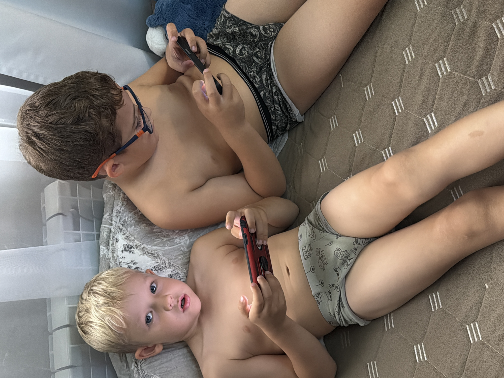 Two young boys are depicted lying shirtless on what appears to be a bed or sofa, both engrossed in using smartphones. The older boy, with brown hair and glasses featuring orange accents, is situated towards the top of the frame, wearing dark grey boxer briefs with a light-colored pattern. He holds a dark smartphone with both hands. A dark blue stuffed animal is partially visible behind his head.
Below him lies a younger boy with blonde hair and blue eyes, wearing light grey briefs adorned with cartoon vehicle patterns. He is looking directly at the camera with wide eyes and a slightly open mouth, momentarily distracted from the red and black smartphone he holds.
The surface they are resting on consists of two different textures: a light grey fabric with a subtle floral or paisley pattern beneath the younger boy's upper body, and a brown quilted bedspread with a hexagonal stitch pattern containing white parallel lines, covering the rest of the area. To the left, a sheer white curtain and a white multi-compartment storage unit are visible, indicating an indoor setting. The ambient light suggests it is daytime. The boys appear focused on their respective devices. No text is visible in the image. The location context for this scene is Sochi, Russia.