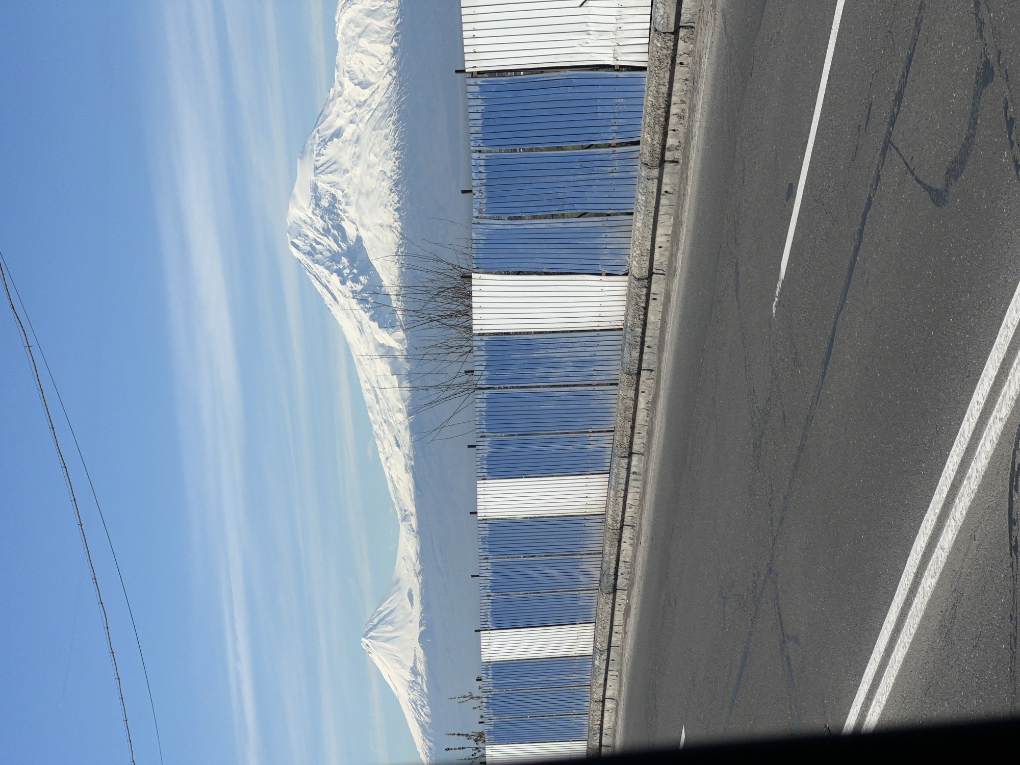 The image shows a roadside view near Yerevan, Armenia. The road runs horizontally across the lower half of the image, marked with white lane lines. A white and blue corrugated metal fence runs alongside the road. Beyond the fence, the snow-capped peaks of Mount Ararat are visible against a blue sky with light, streaky clouds. Bare tree branches are visible in front of the fence. A power line stretches across the upper left portion of the sky.