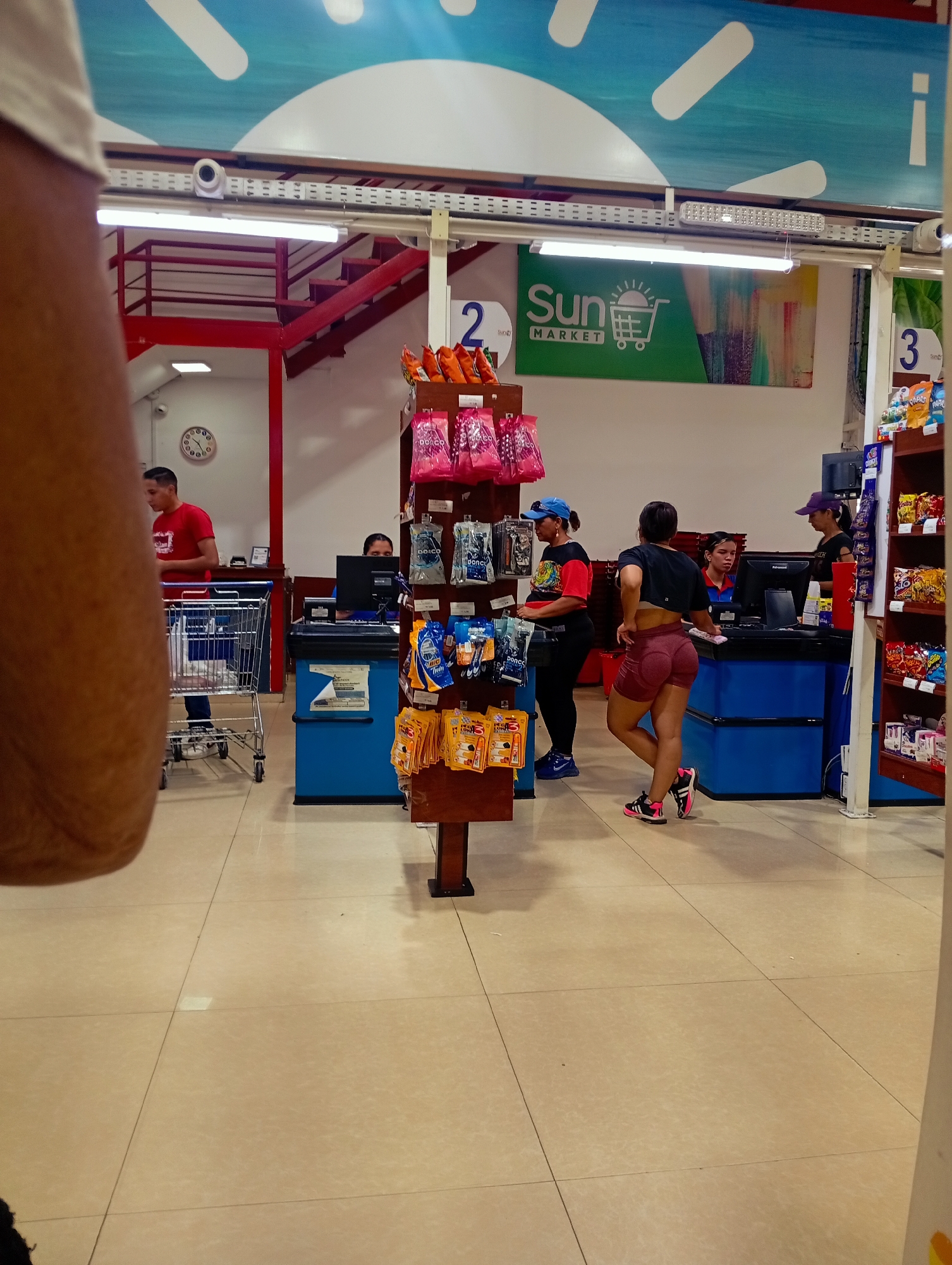 The image shows the interior of a "Sun Market" store in Juan Griego, Venezuela. The focus is on the checkout area, where three cashiers are stationed behind blue counters. Two female customers are present; one is wearing a blue cap and the other is wearing a black cropped top and red shorts. A display of small, packaged goods stands in front of one of the checkout lanes. A man with a shopping cart waits at a different checkout lane in the background. Red stairs and a clock are visible in the background. The floor is tiled and the store appears to be brightly lit with overhead fluorescent lights.