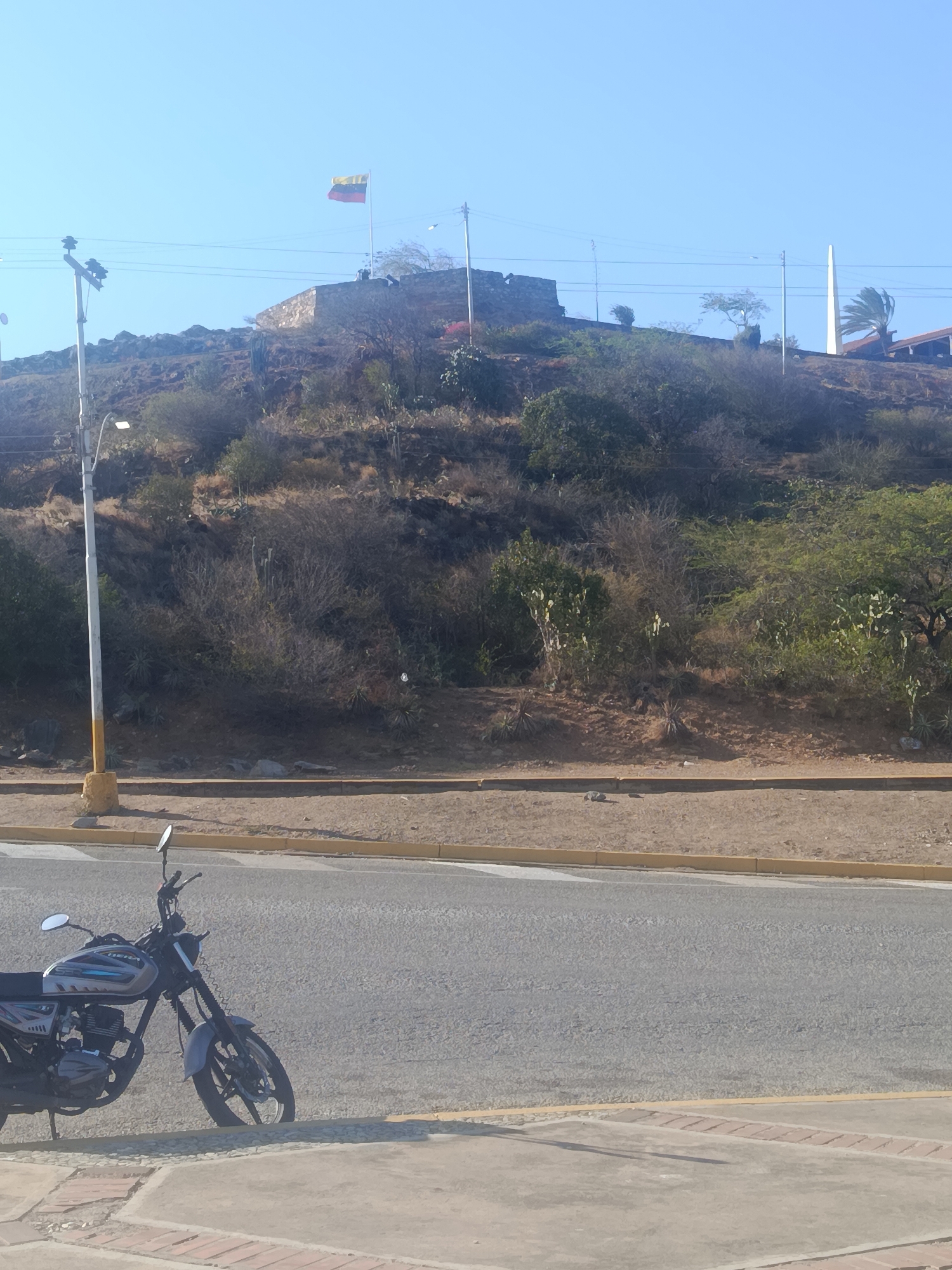 The image shows a street scene in Juan Griego, Venezuela. A motorcycle is parked on the sidewalk beside a road. Beyond the road, a hill rises up with sparse vegetation and a stone structure with the Venezuelan flag flying on top. There's also a tall, white obelisk visible on the hillside. A street lamp stands near the sidewalk, with power lines visible in the sky. The overall impression is of a sunny, somewhat arid environment.