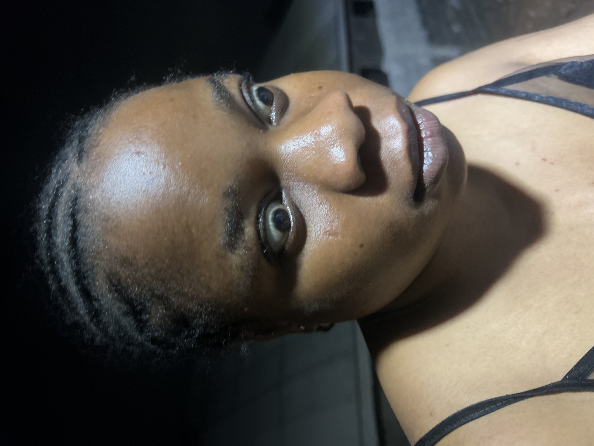 A close-up photograph depicts an individual with dark skin, facing slightly to the right and upward. The person's hair is styled in dark braids that frame the forehead. Their eyes are open, and their facial skin appears moist. The person is wearing a dark, thin-strapped garment, revealing part of the shoulder and chest. The background is dark, with a faint, indeterminate shape visible in the upper left corner. The location in the city is Unknown, Unknown.