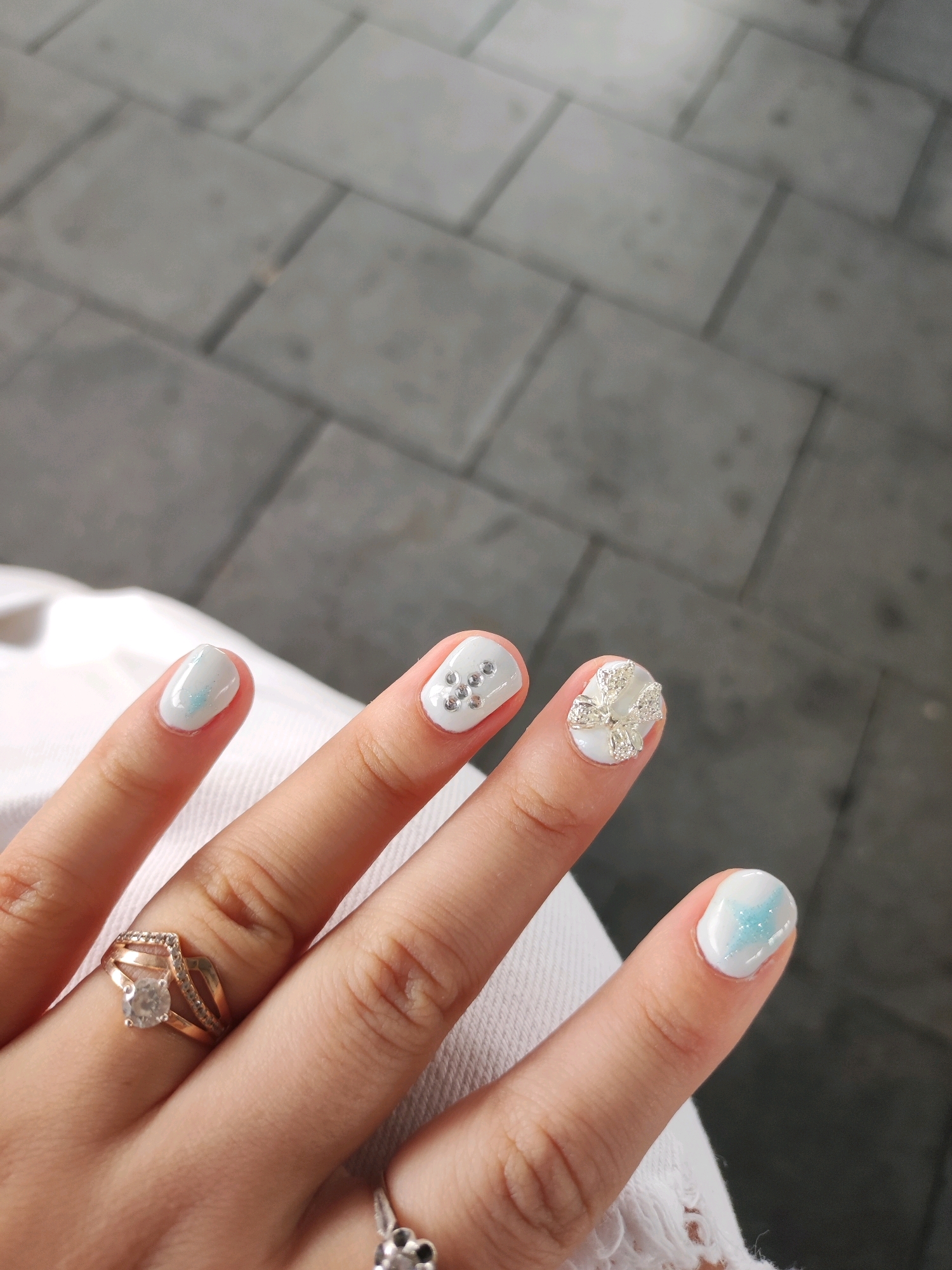 The image shows a close-up of a person's hand with a manicure and rings. The nails are painted white with some glitter and blue accents on the thumb and index finger. The middle finger has small silver rhinestones, and the ring finger has a 3D bow embellishment with rhinestones. The person is wearing a gold ring with a clear stone on the ring finger and another ring with a flower design on the pinky finger. The background appears to be an outdoor tiled surface, likely a walkway or patio. The image is taken from a slightly overhead perspective, focusing on the hand. There is no information in the image to indicate it is located in Ansbach, Germany.