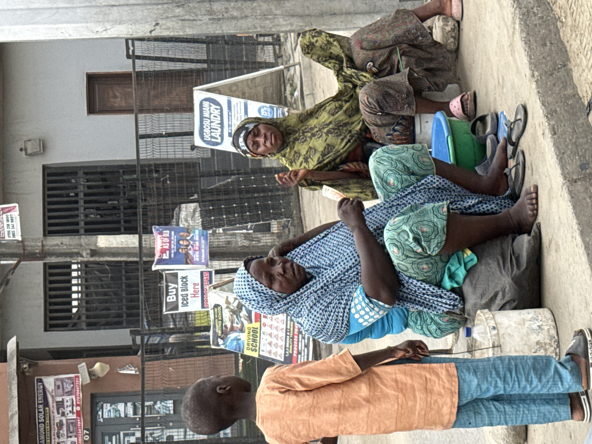 This media file captures an outdoor street scene in Lekki, Nigeria, featuring three individuals and several commercial signs. The image is taken during bright daylight, suggesting daytime.
In the foreground, a young boy with a shaven head stands with his back to the viewer. He is wearing an orange long-sleeved top, blue trousers, and black and white Adidas sandals. He appears to be facing two adult women who are seated on the ground or a low ledge beside a building.
The woman closer to the viewer (left-center) wears a blue and white patterned head covering and a light blue and green patterned outfit. She looks towards the camera with a neutral expression. Next to her are a blue plastic bucket, a green plastic basin, and several pairs of flip-flops. The woman behind her (right-center) is dressed in a green patterned head covering and a brownish patterned dress, also looking towards the camera. A grey bag or cushion serves as her support. Scattered around them are more flip-flops and a white bucket with a plastic bottle.
The background consists of a building with several barred windows and a brown wooden panel. The walls are adorned with various advertising signs. Visible text includes "UGBOSU MIAMI LAUNDRY" (appearing twice), "DRIVING SCHOOL" (also twice, one mentioning "QUALITY TRAIN" and "1,2,3 WEEKS & MONTH"), "Buy Iced Block Here," and "AS OF OLD." Another sign advertises "DIAMOND SOLAR ENERGY - POWER OF LIGHT." Several phone numbers are also displayed: "0705 218 1800," "0803 341 9897," "08033419897," "07070447351," and "09040637615." A "15%" discount is visible on one of the laundry signs. The overall scene depicts a typical, bustling urban setting.