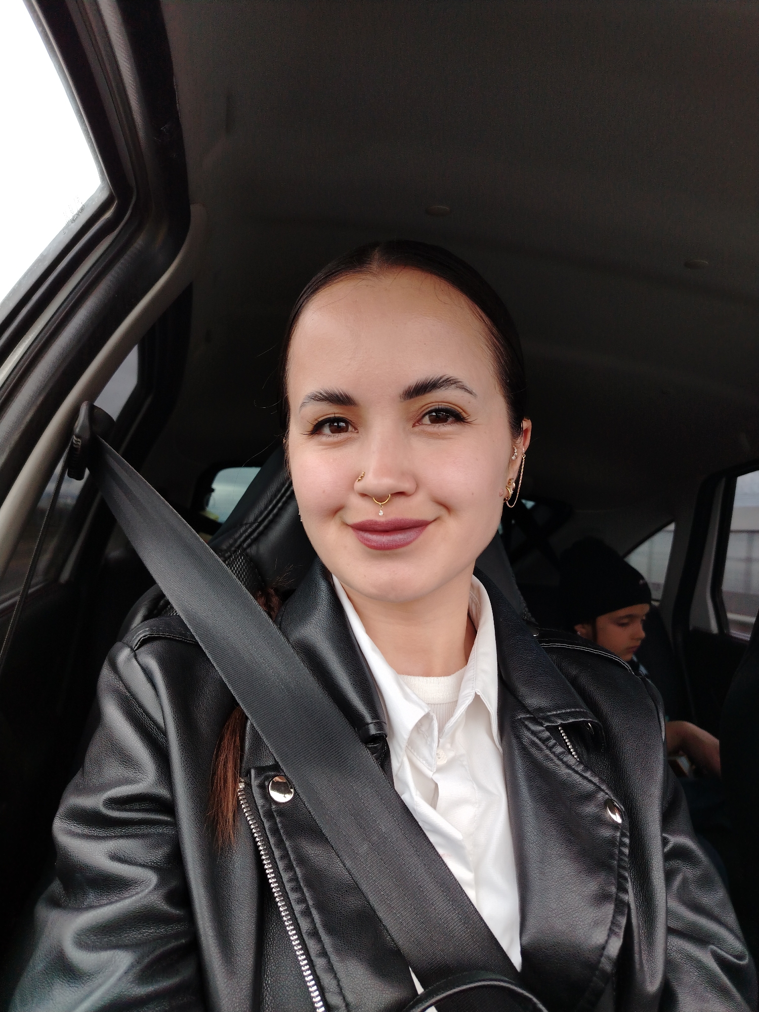 This image captures the interior of a vehicle located in the city of Chaikovsky, Russia. The primary subject is a female adult seated in the front passenger seat, secured by a black seatbelt. She has dark hair pulled back, brown eyes, and is looking directly at the camera with a subtle smile. Her facial features include a gold-colored septum ring with a small clear gem, a small gold stud on her right nostril, and multiple gold-colored earrings on her right ear, including a chain connecting two piercings. She is wearing dark red or plum-colored lipstick. Her attire consists of a black faux leather jacket over a white collared shirt, beneath which a white ribbed garment is visible.
In the background, a second individual, a child or young adolescent, is visible in the rear passenger seat on the right side of the vehicle. This individual is wearing a black beanie hat and a dark-colored top, and their face is seen in profile, looking downwards or away from the camera.
The vehicle's interior features black leather or faux leather seats and a dark gray headliner. Through the front left window, a bright, possibly overcast sky is visible. The view through the rear passenger window is blurred and indistinct, suggesting the car may be in motion or parked near an undifferentiated background.