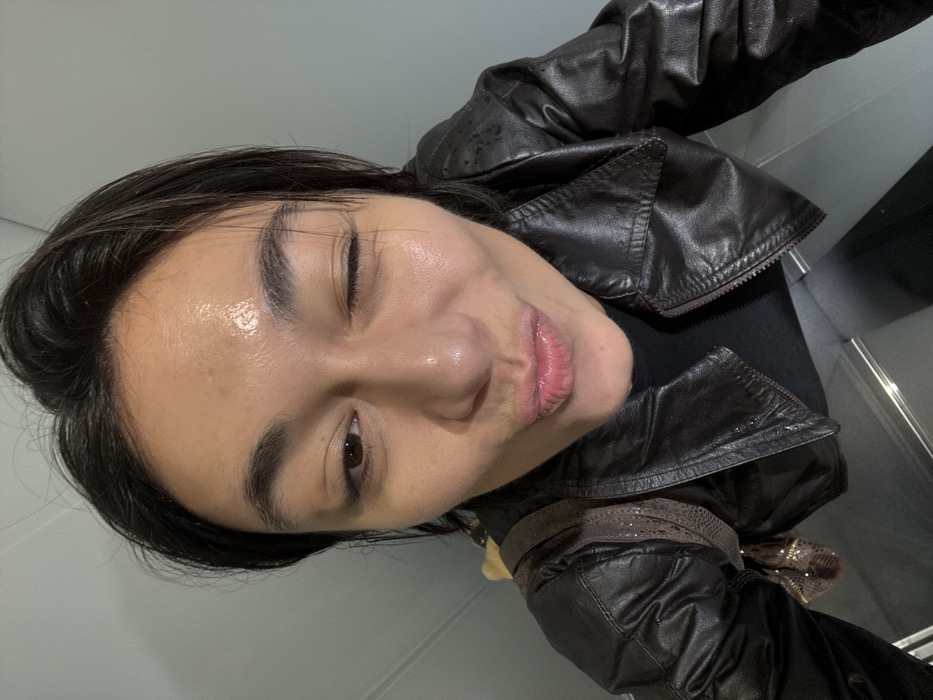 The image presents a close-up, high-angle view of a female individual indoors in Paris, France. Her complexion is fair, displaying a noticeable sheen across the forehead, nose, and cheeks. She is depicted with her left eye closed in a wink and her lips puckered. Attire includes a dark brown leather jacket over a black top. A portion of a brown shoulder bag with a patterned texture, possibly snakeskin, is visible over her left shoulder. The immediate background is composed of smooth grey panels, transitioning to a darker horizontal surface at the bottom with a distinct reflective line.