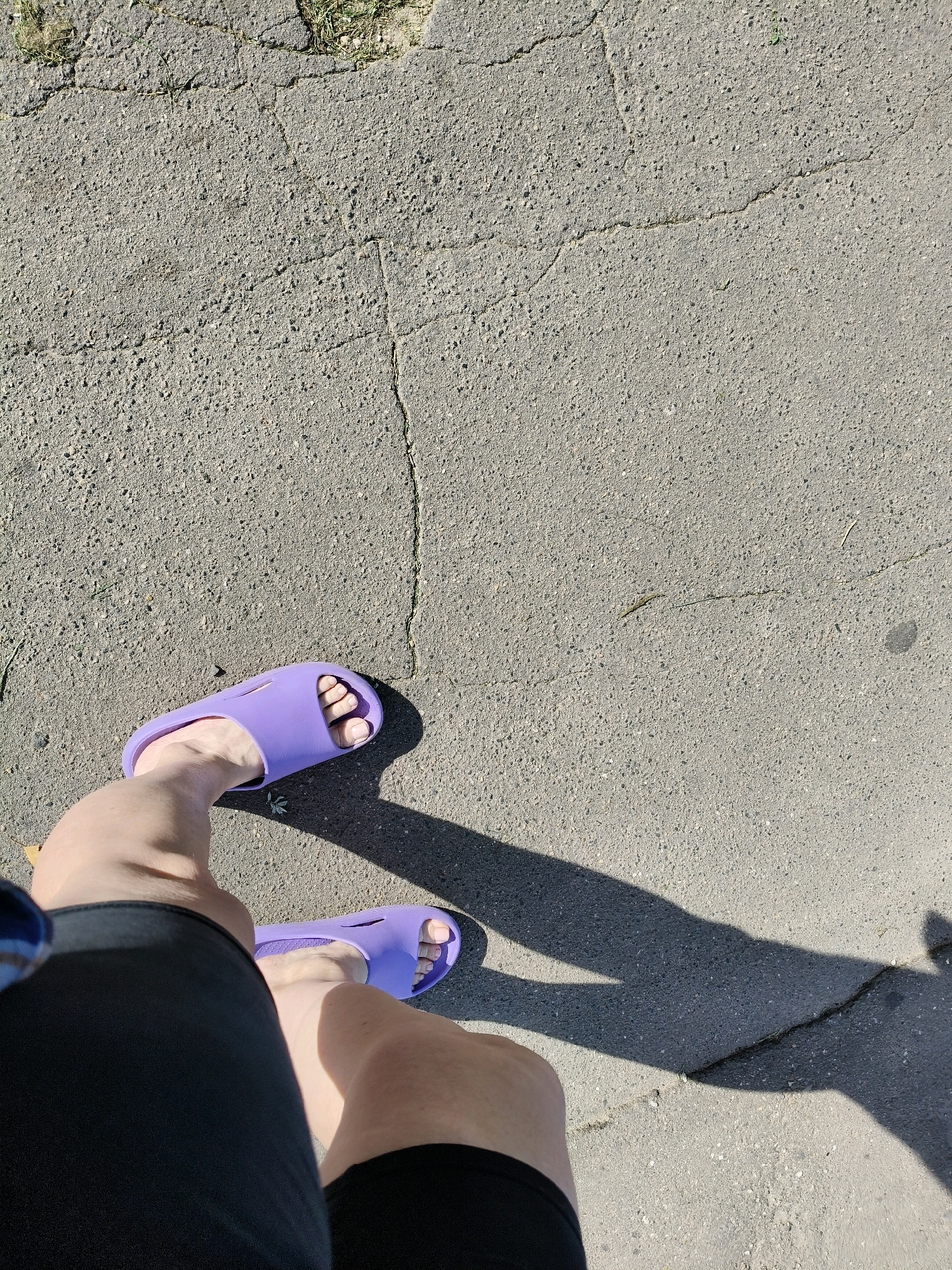 A high-angle shot shows a person's legs and feet, wearing purple slides and black shorts, standing on a cracked asphalt surface. A shadow is cast by the person and their footwear. The scene appears to be outdoors on a sunny day.