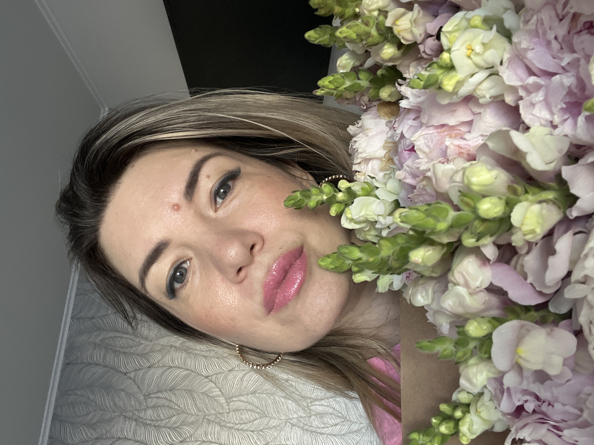 A woman with blonde hair, wearing a pink shirt, is holding a bouquet of pink and white flowers. She has on eye makeup and pink lipstick, and she is wearing a hoop earring with small diamonds. A small mole is visible on her forehead. The background is a textured wallpaper and a gray wall, suggesting an indoor setting. The lighting suggests it could be daytime. The image appears to be a selfie.