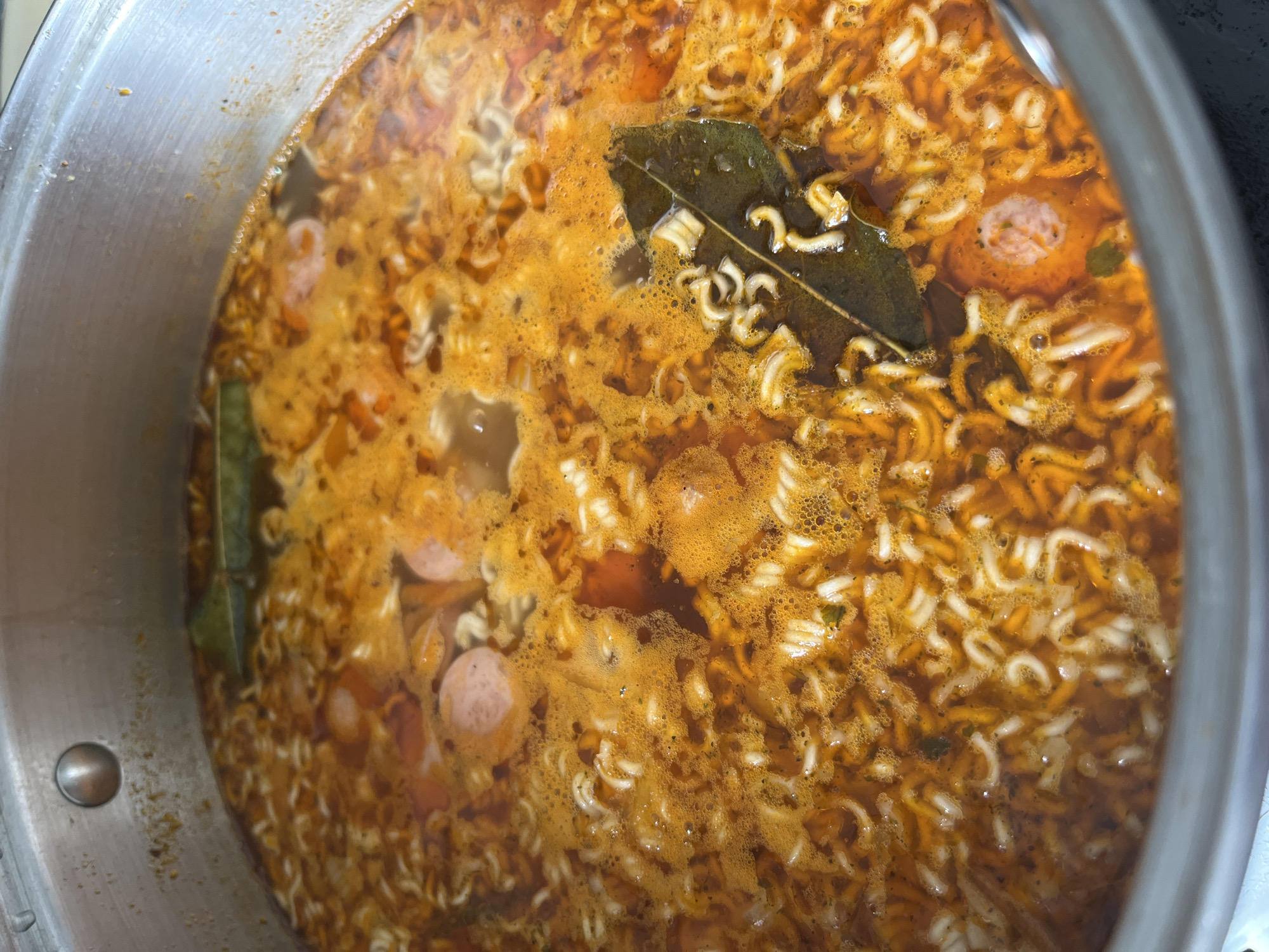 The image is a close-up, overhead shot of a pot of instant noodles cooking in a broth. The noodles are curly and pale white, submerged in a reddish-orange, frothy broth. Several slices of pink sausage are visible amongst the noodles. Two large, dark green bay leaves are floating on the surface of the broth. The pot appears to be made of brushed metal, with some slight discoloration and residue visible on the rim. The background is dark and out of focus. There is no information available in the image to determine its location in Chelyabinsk, Russia.