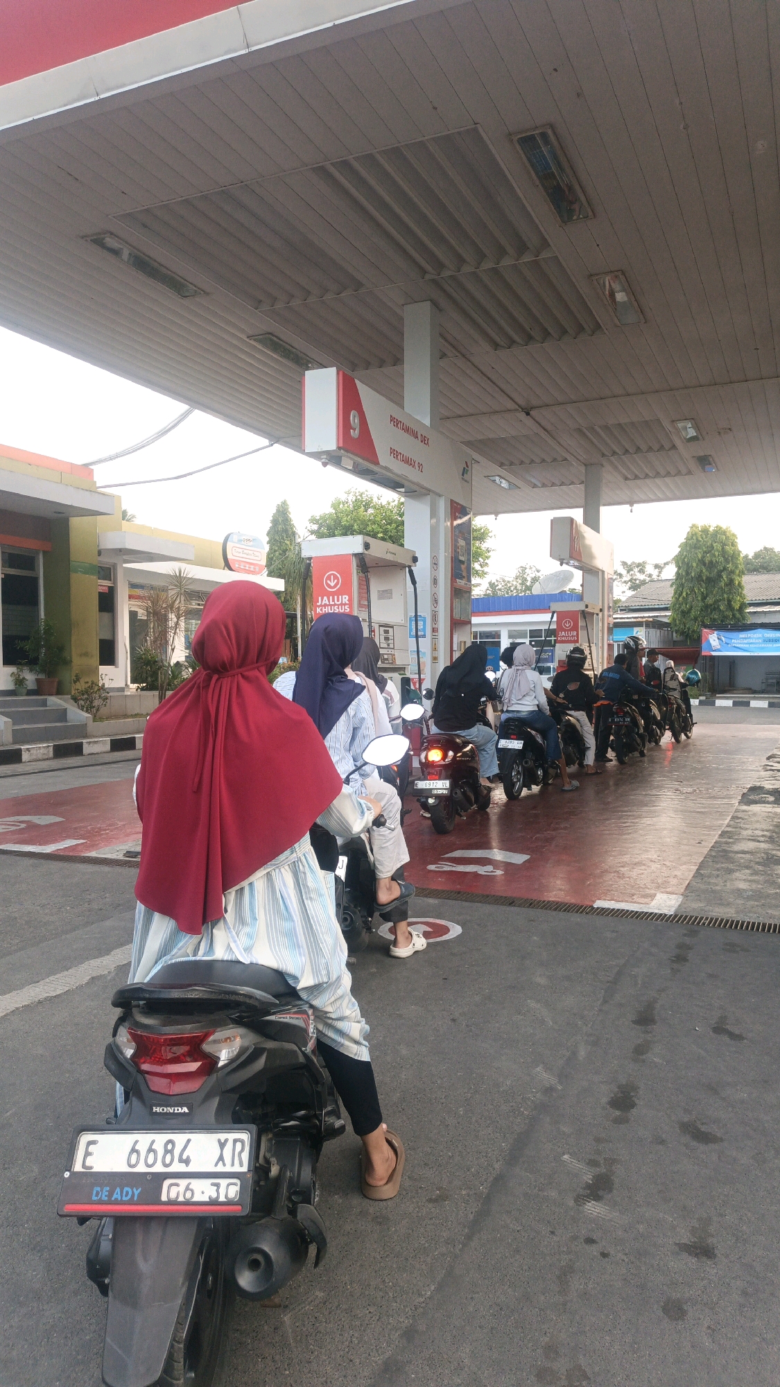 The image depicts a queue of motorcyclists at a Pertamina gas station in Cipinang, Indonesia. In the foreground, a person wearing a red hijab and a striped shirt is seated on a black Honda motorcycle, facing away from the camera. The motorcycle's license plate reads "E 6684 XR DE ADY G6.30". Several other motorcyclists, mostly wearing hijabs and long sleeves, are lined up behind the first motorcycle, waiting to refuel at the gas pumps. The gas station has a covered canopy supported by white pillars. Signs on the canopy indicate "PERTAMAX DEX" and "PERTAMAX 92". The ground is paved and marked with red lines. In the background, there are buildings associated with the gas station and some trees. The overall scene suggests a typical day at a gas station with customers waiting in line.