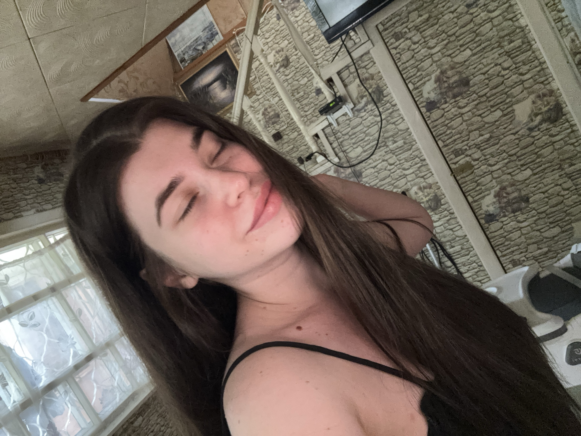 A young woman with long, dark hair is seen in a tilted close-up shot with her eyes closed. She is wearing a black, thin-strapped top. A mole is visible on her neck. In the background, there is a wall with a patterned brick-like design. A television and framed pictures are mounted on the wall. To the left, a window is visible with a sheer curtain. The location appears to be indoors. There is no evidence to identify this location is in the city of Мещёры, Russia.