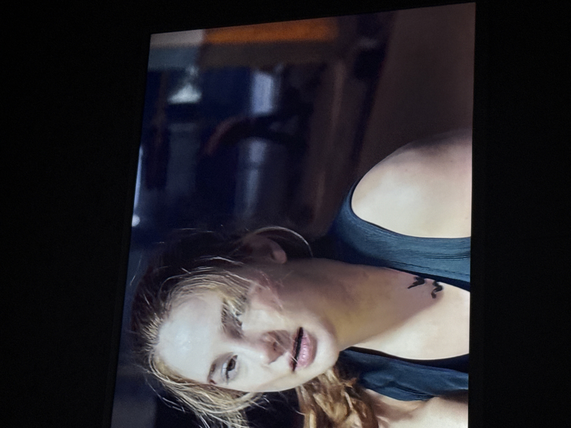 A close-up shot of a blonde woman with a tattoo on her upper chest. She is wearing a tank top. The background is blurry, with hints of industrial or gym equipment. There's no information available to discern the specific location in Mytishchi, Russia.