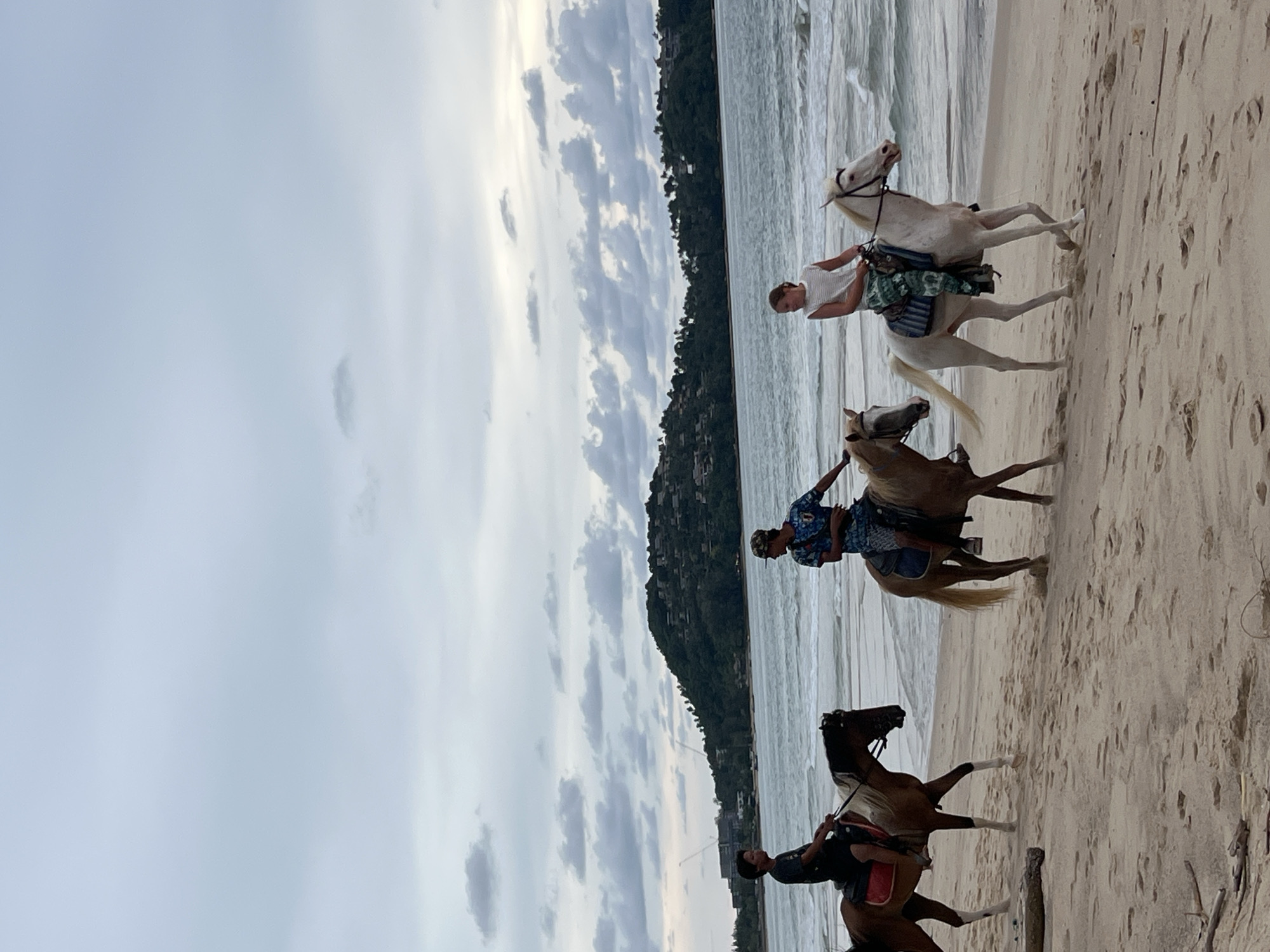 Three people are riding horses on a sandy beach. The person in the front is riding a white horse. They are wearing a white shirt and a patterned skirt. The second person is riding a light brown horse and is wearing a blue and white patterned shirt and blue shorts. The third person is riding a darker brown horse and is wearing a dark t-shirt. They are all facing towards the right of the image. The sea is visible in the background, with waves rolling onto the shore. A forested hill with some buildings is visible behind the sea. The sky is overcast with clouds. The scene appears to be set in the late afternoon or early evening. The overall mood is peaceful and relaxed. There is no visible text in the image.