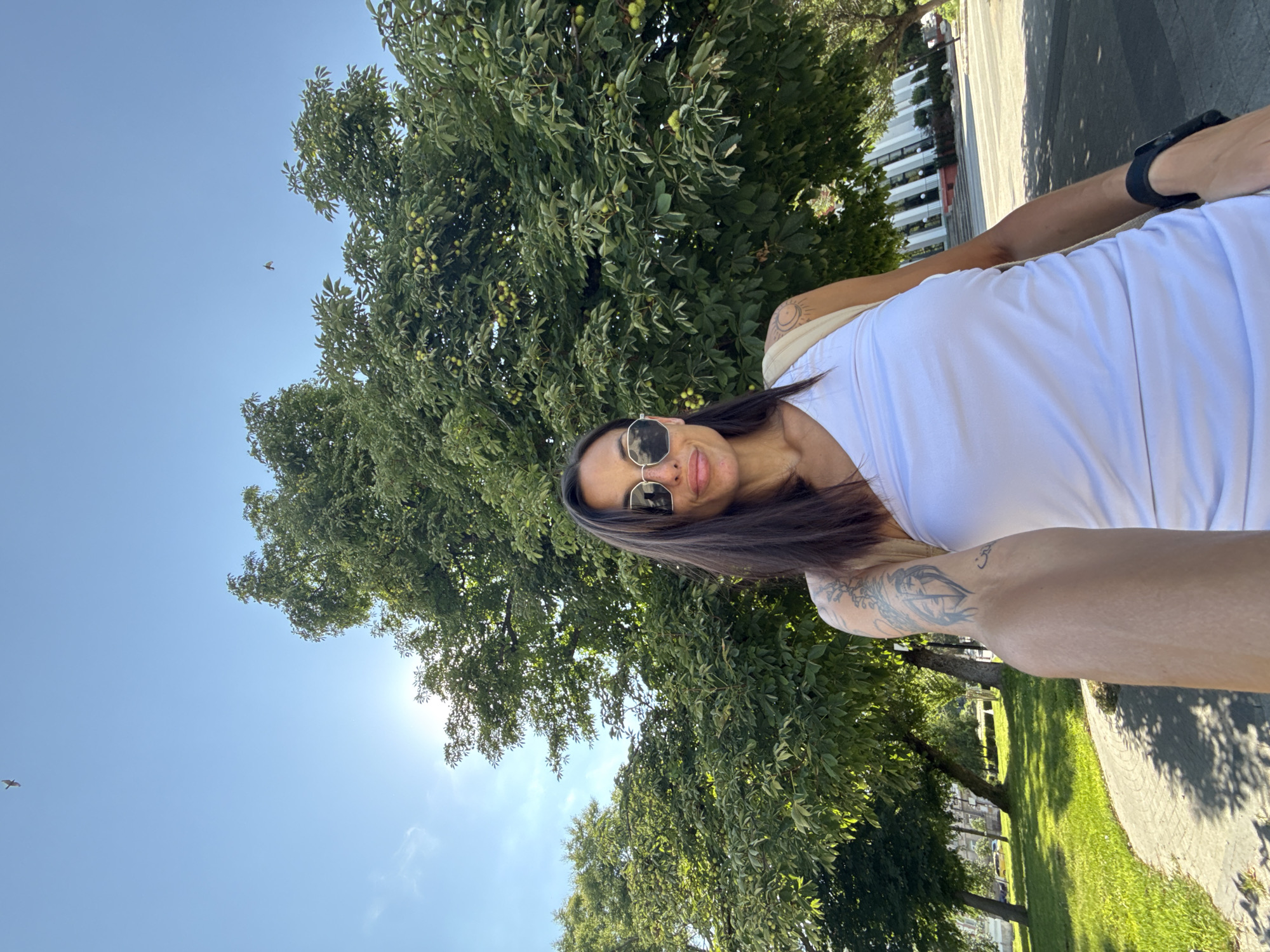 A woman, facing forward and holding a phone for a selfie, stands in a park. She has long dark hair, sunglasses, and is wearing a white tank top. She has tattoos on her arm. Behind her, a large tree with green leaves and some green fruit is visible. The sky is blue with some clouds. The ground is paved with a sidewalk and grass to the left. In the background, there are more trees and a building. The location is identified as the park near the Pleven Epopee.