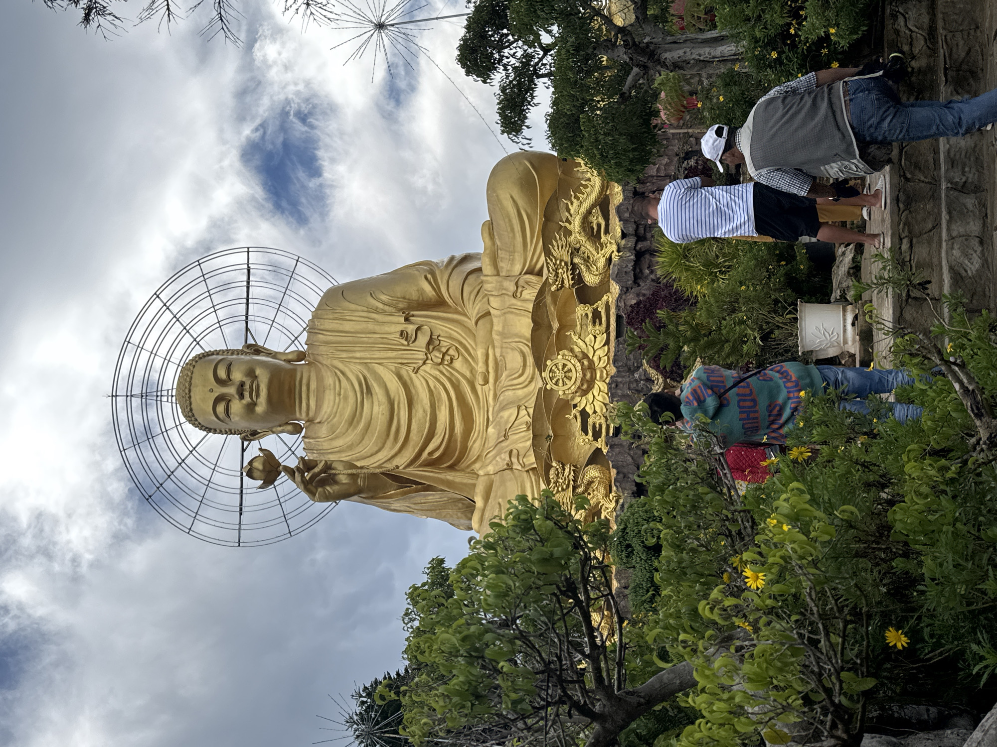 In Lâm Viên - Đà Lạt, Vietnam, a colossal golden Buddha statue sits serenely against a cloudy sky, holding a lotus flower in its right hand. The statue is adorned with a circular halo and rests on a pedestal featuring intricate golden dragon sculptures. In the foreground, a group of people are visible, some walking up stone steps and others observing the monument. Lush greenery, including trees and yellow flowers, surrounds the base of the statue and frames the scene, with a stone wall forming the backdrop. Two individuals are captured from behind, one wearing a striped t-shirt and shorts, the other a vest over a plaid shirt. Another person, facing away, is dressed in a hooded sweatshirt and jeans.