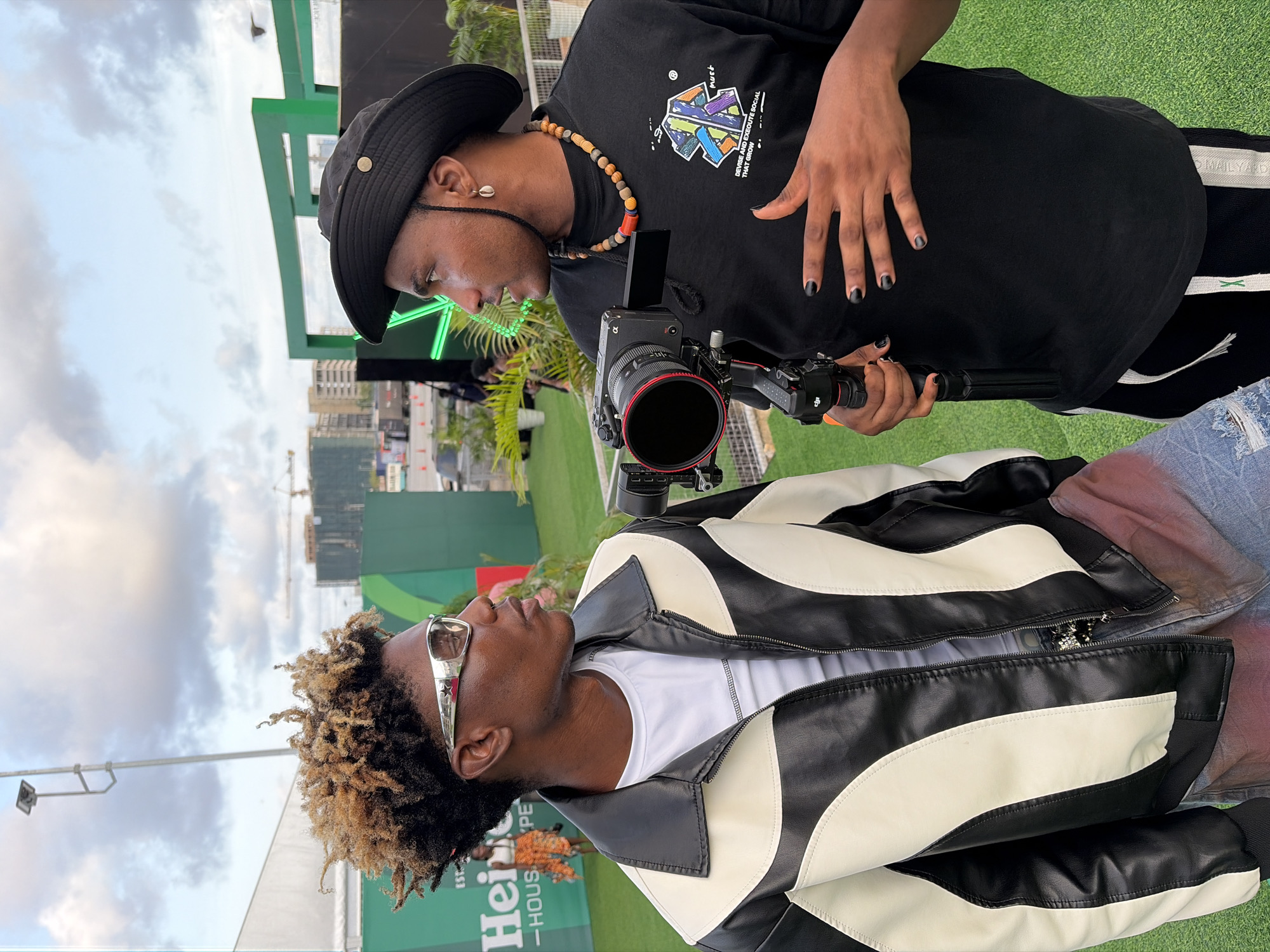 Two African men are standing face to face in an outdoor setting that appears to be in Lagos, Nigeria, indicated by the urban skyline in the background and the Heineken signage. The man on the left has light brown dreadlocks, sunglasses with a star design, a white t-shirt and a black and white leather jacket. The man on the right is wearing a black bucket hat, a beaded necklace, and a black t-shirt with a colorful graphic design. His nails are painted black. He is holding a professional-grade camera with a long lens and an attached stabilizing device. The men are engaged in conversation. The background includes green artificial turf and buildings.