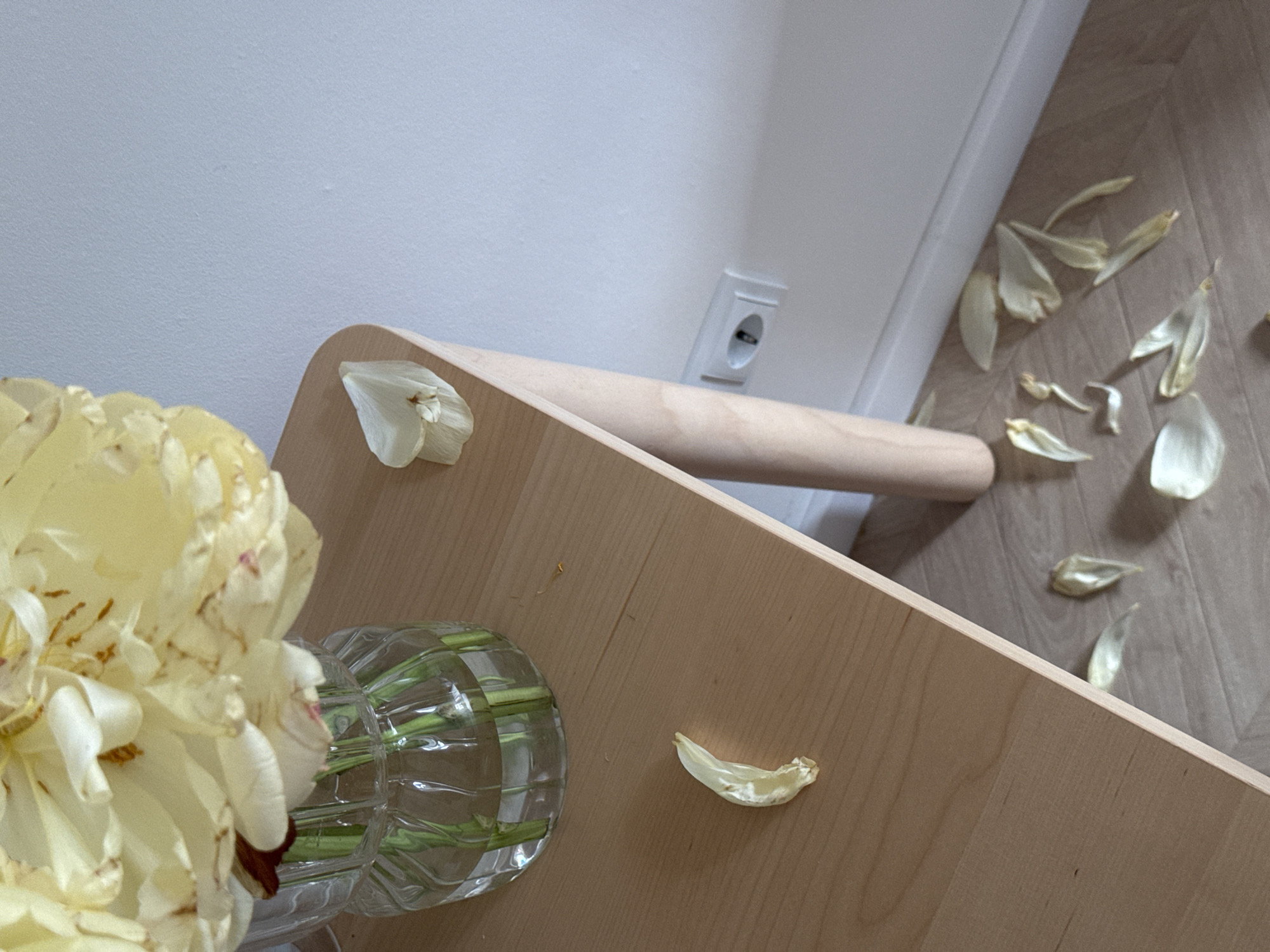 The image shows a still life composition. A light yellow flower arrangement in a clear glass vase sits on a light-colored wooden surface. The surface appears to be part of a small table or platform. Several fallen flower petals are scattered on the table and the floor, which has a light wood grain pattern. In the background, a plain white wall is visible with an electrical outlet. The angle of the shot is from above, looking down and slightly to the side. The location is unknown, and no information suggests it is in Люк, Russia.