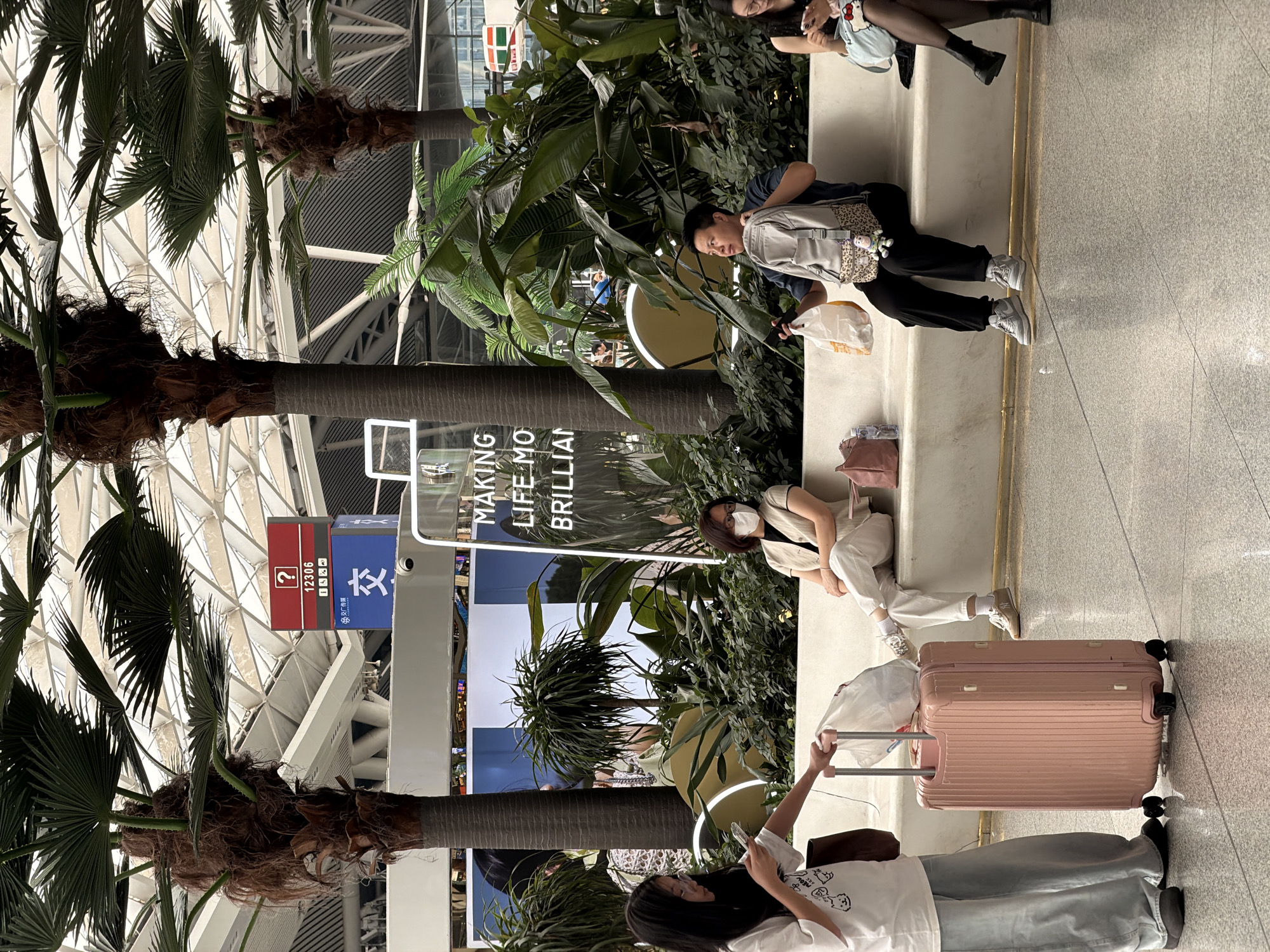 A group of people are waiting in a public indoor space that resembles a transportation hub or large waiting area, possibly Panyu District, China, due to the presence of Chinese signage and architectural style. The environment features large palm trees and abundant greenery, with a modern, light-filled ceiling structure.
In the foreground, a young woman wearing glasses, a white t-shirt with a graphic print, and baggy light blue pants stands holding a pink suitcase. She is looking at her phone. Next to her, a woman in a light beige outfit with a face mask is seated on a white bench, looking forward. To her right, a man wearing a dark shirt, black pants, and gray sneakers is seated, holding a white paper bag in his right hand and a gray backpack with a leopard print strap and a small figurine attached in his left. He is looking up and to the right. Further to the right, a woman with dark hair is seated on the bench, wearing a black top and dark patterned tights, looking down at her phone.
In the background, signage is visible, including a red sign with a question mark and a number "12306," and a blue sign with Chinese characters, likely indicating transportation information. A neon sign with text "MAKING LIFE MO BRILLIAN" is also visible. The floor is tiled with a reflective surface.