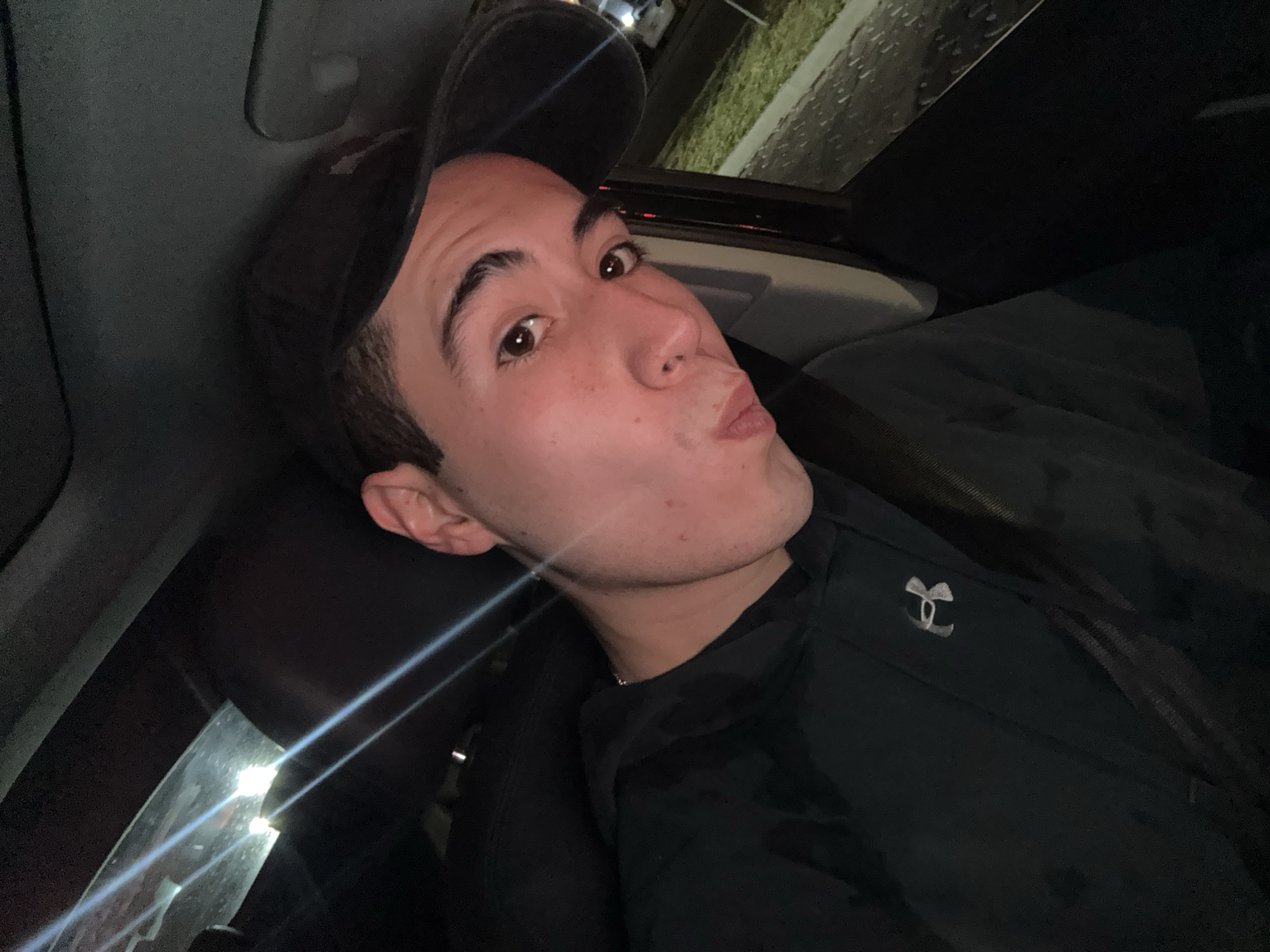 A single male individual is depicted in a close-up, angled portrait, presumably a selfie, taken inside a vehicle at night. The specific location within Heroica Puebla de Zaragoza, Mexico, cannot be determined from the visual evidence.
The subject is a young male with fair skin, dark hair, dark eyebrows, and brown eyes. He exhibits reddish spots or freckles on his cheeks and nose. He wears a dark baseball cap, backward, and a dark hooded sweatshirt displaying a silver "Under Armour" logo on the left chest. A thin, light-colored chain is visible around his neck. A dark seatbelt strap crosses diagonally over his chest and left shoulder. The individual is looking directly at the camera, with his lips pursed in a "kissing" expression.
The interior environment appears to be the passenger cabin of an automobile, characterized by dark upholstery and surfaces, including a dark headliner. A strong, bright light source in the upper left creates a pronounced lens flare. Through the right-side window, an exterior nighttime scene is visible, including a strip of green grass, a paved or curbed area, and indistinct distant lights, suggesting a street or parking lot environment.