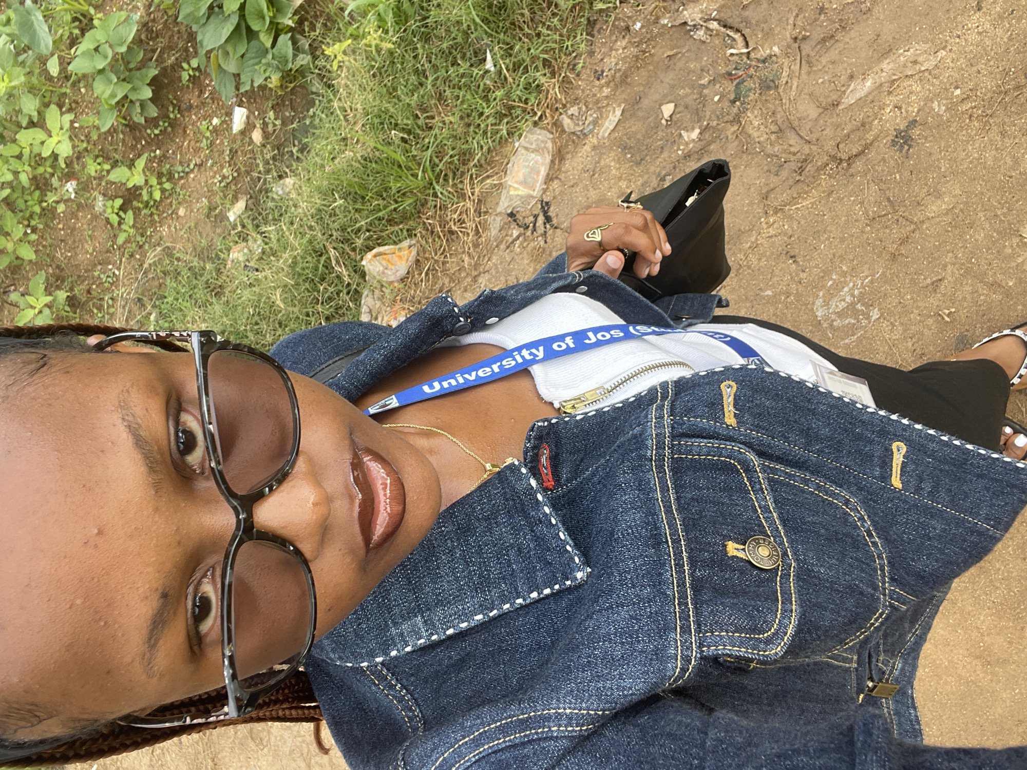 A young Black woman with braided hair, wearing large, dark-rimmed glasses, a denim jacket over a white top, and dark pants, stands outdoors looking at the camera. A blue lanyard with "University of Jos" printed on it hangs around her neck. She is holding a black bag in her right hand and wearing rings on her fingers. The ground is dirt and gravel, with patches of grass and scattered debris, including plastic bags. The scene appears to be in a semi-urban or rural outdoor environment, possibly in Rafin Par, Nigeria.