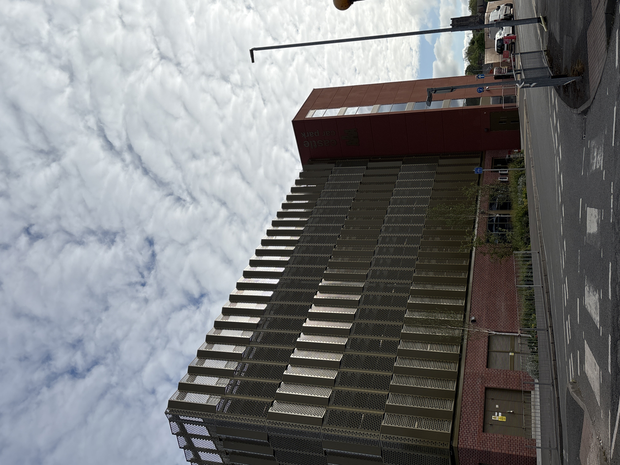 The image captures the Castle car park in Newcastle-under-Lyme, United Kingdom, from a low-angle perspective. The car park's distinctive stepped facade of a metal perforated screen dominates the foreground, adjacent to a red brick section with two closed service doors. Above this is a rust-colored, rectangular building extension with a horizontal row of windows along the top. The building has signage that is difficult to read, appearing to say "Castle Car Park."
A street runs parallel to the building. Several parked cars and vans are visible in the background. Street furniture includes a tall streetlight, pedestrian barriers, and signs.
The sky is heavily clouded, suggesting overcast weather.