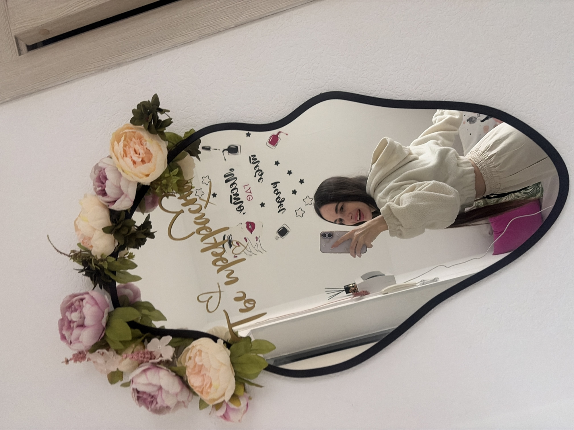 A woman is taking a selfie in a heart-shaped mirror that is decorated with flowers. The mirror has the phrase "Be Perfectly You" written on it, along with beauty-related images. The reflection shows a white shelf with objects and a pink pillow in the background. The mirror is mounted on a white wall. The image does not reveal any information to indicate that the location is Saint Petersburg, Russia.
