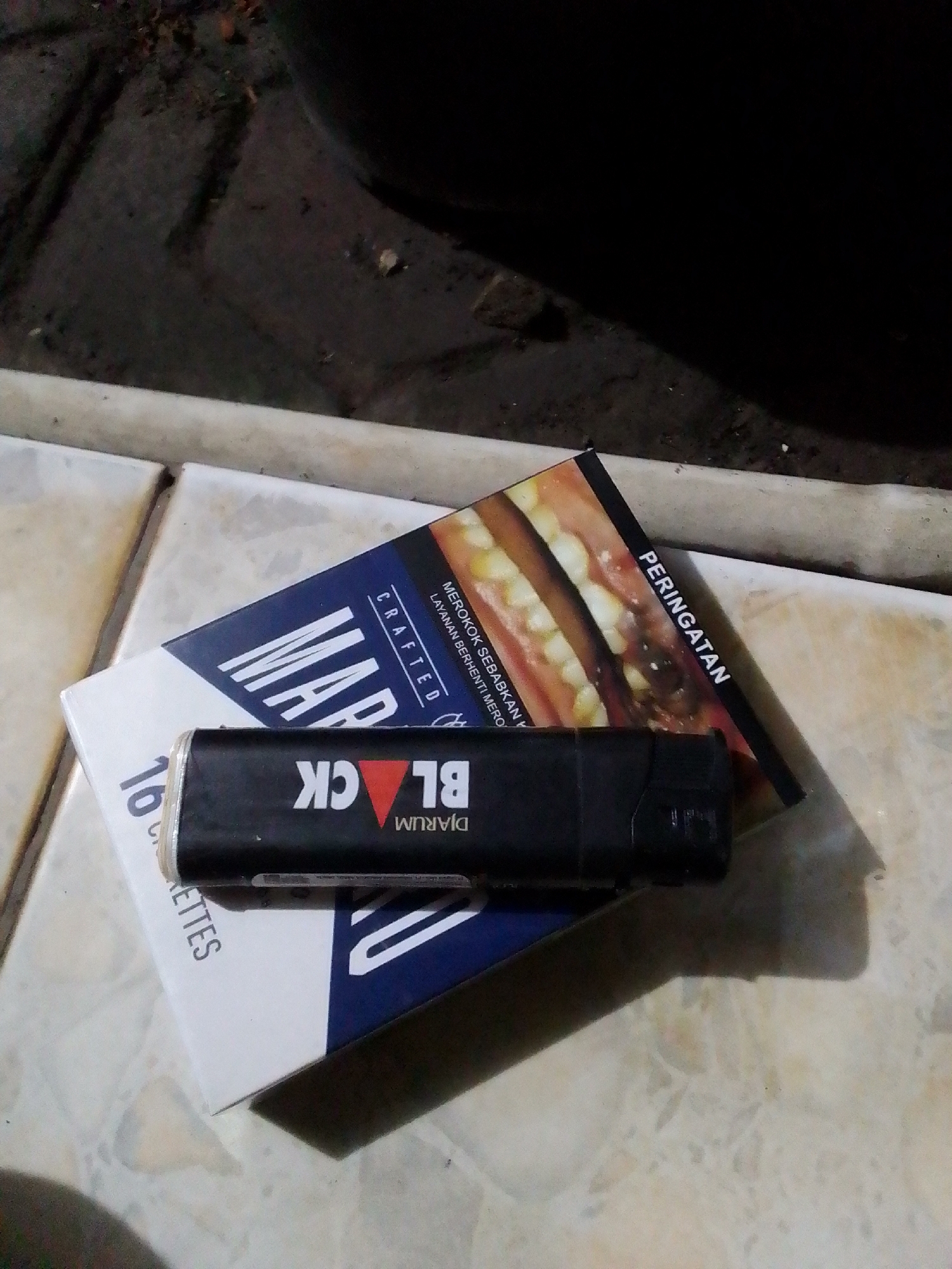 A black cigarette lighter with the words "DJARUM BLACK" printed on it rests on top of a pack of Marlboro cigarettes. The Marlboro pack is partially obscured by a cigarette pack with a warning label. The items are on a tiled surface. The image appears to be taken outdoors at night, possibly in an urban environment. The background is dark and out of focus, with some debris visible on the ground.
